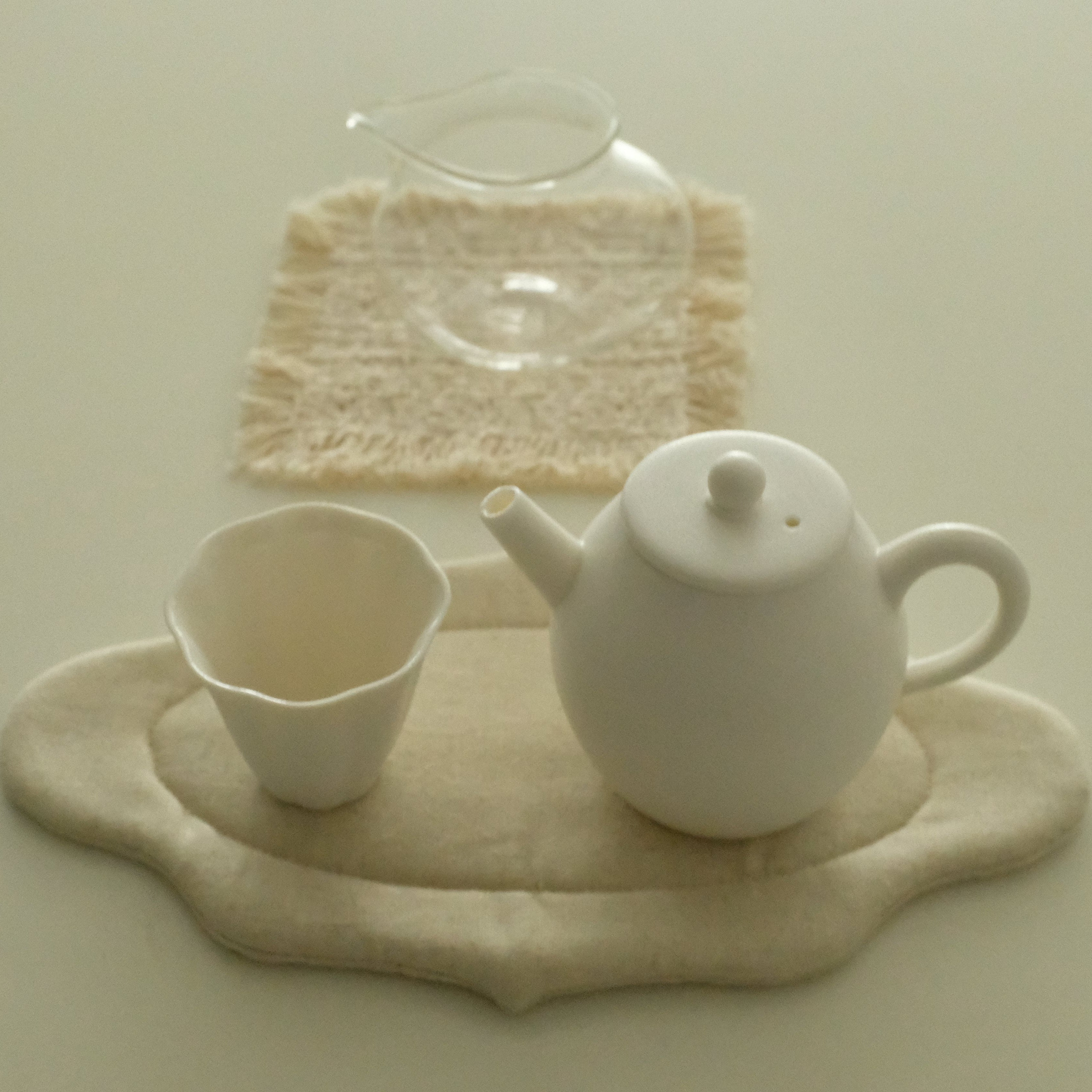 A small white ceramic teapot and matching cup arranged on a beige fabric mat, with a clear glass pitcher resting on a textured cream cloth in the background.