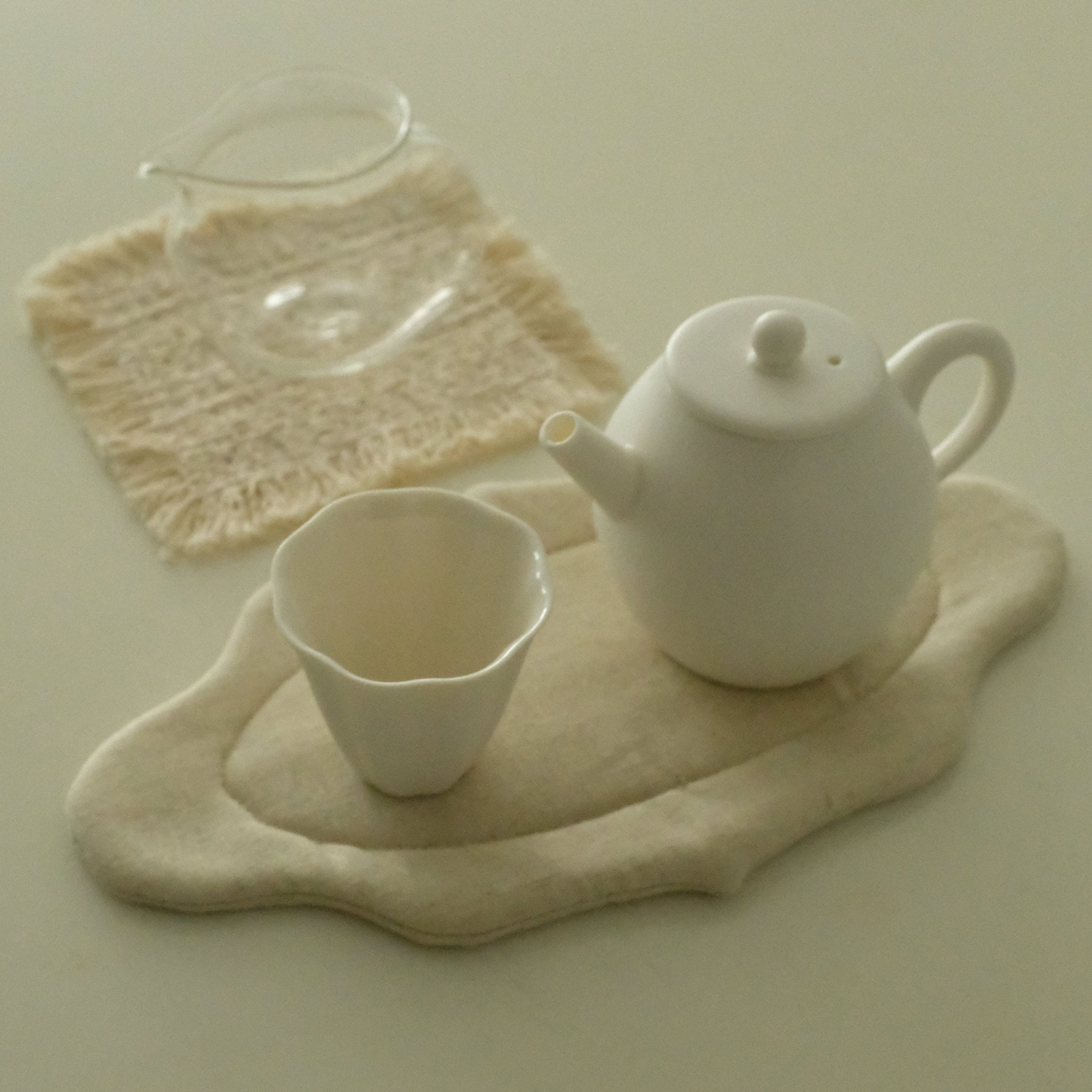 A white ceramic teapot and matching cup arranged on a cream fabric tea mat, with a small clear glass pitcher on a lace cloth behind it, all on a plain light beige background.