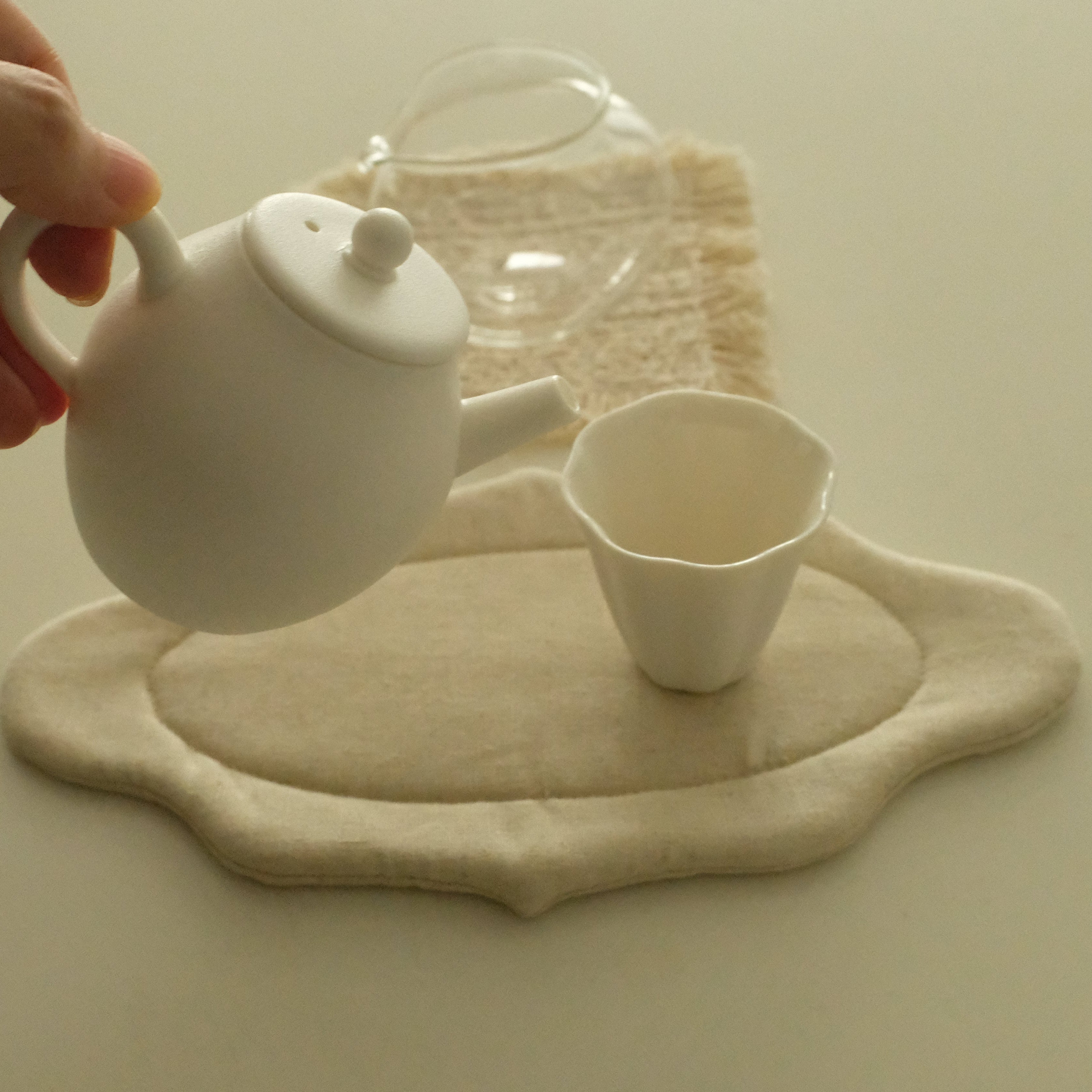 A hand holds a small white teapot above a matching white cup and a beige fabric tea mat on a light neutral surface.