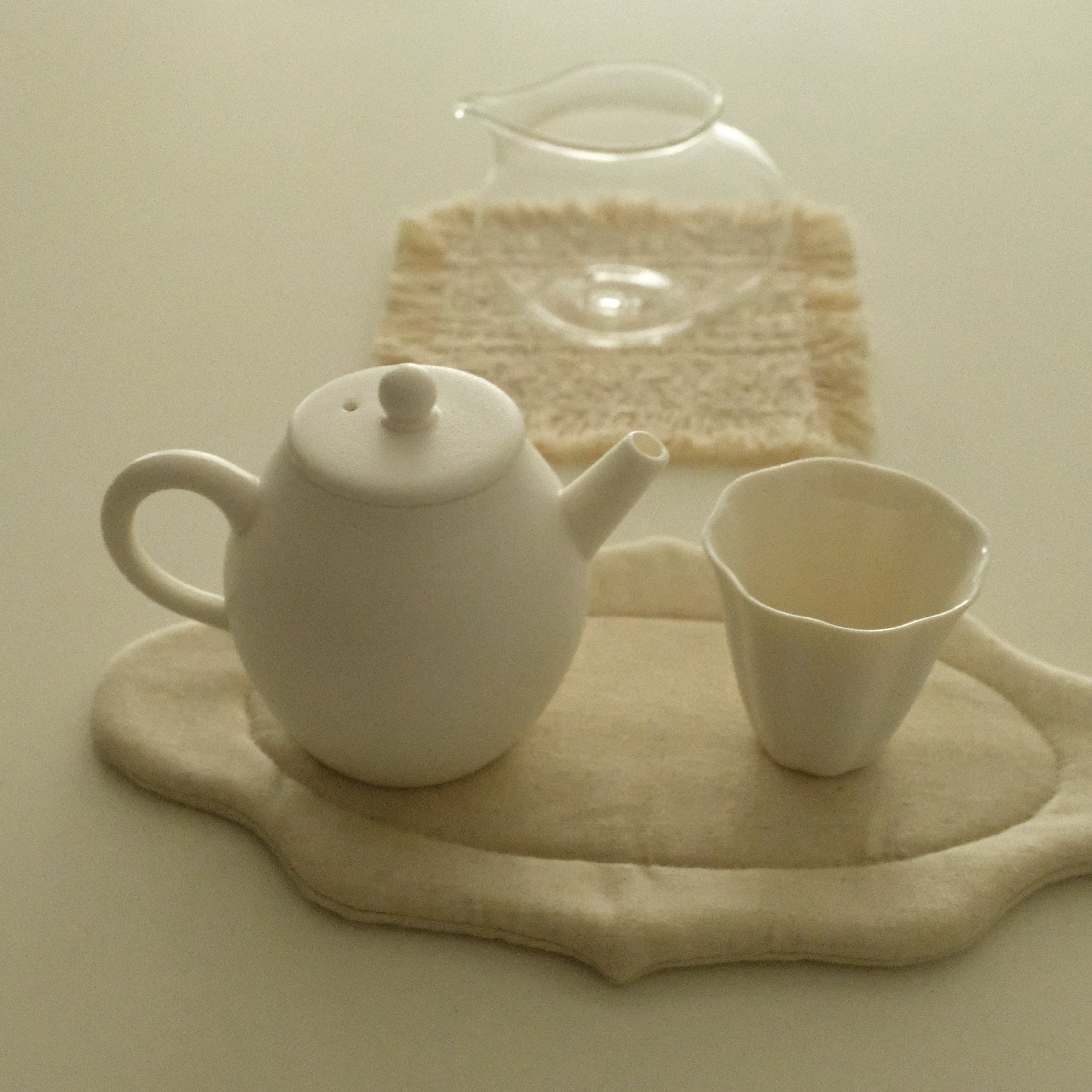 A white ceramic teapot and matching cup arranged on a beige fabric tray, with a small clear glass pitcher resting on a textured beige cloth in the background.