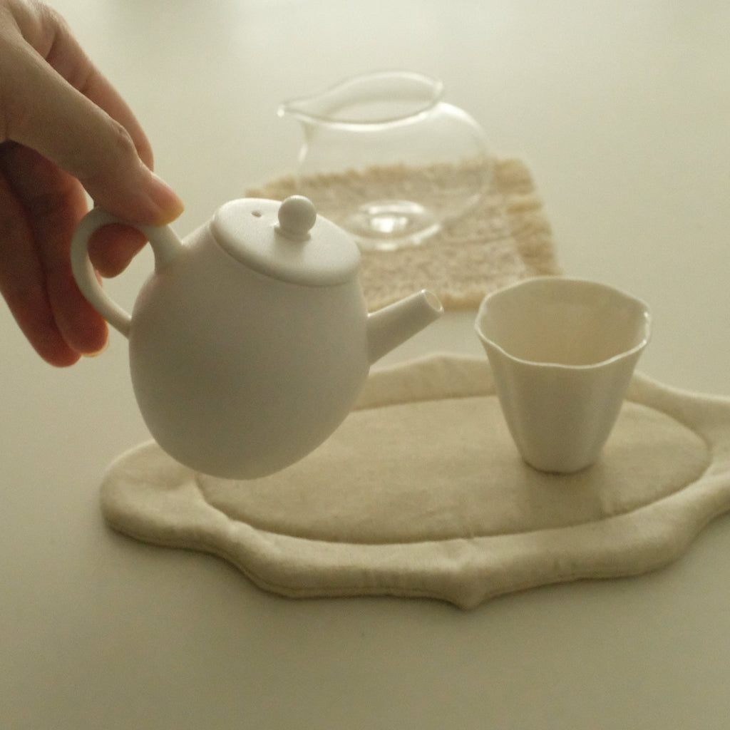 A hand holds a small white ceramic teapot above a matching white cup and a beige fabric tea mat on a light surface.