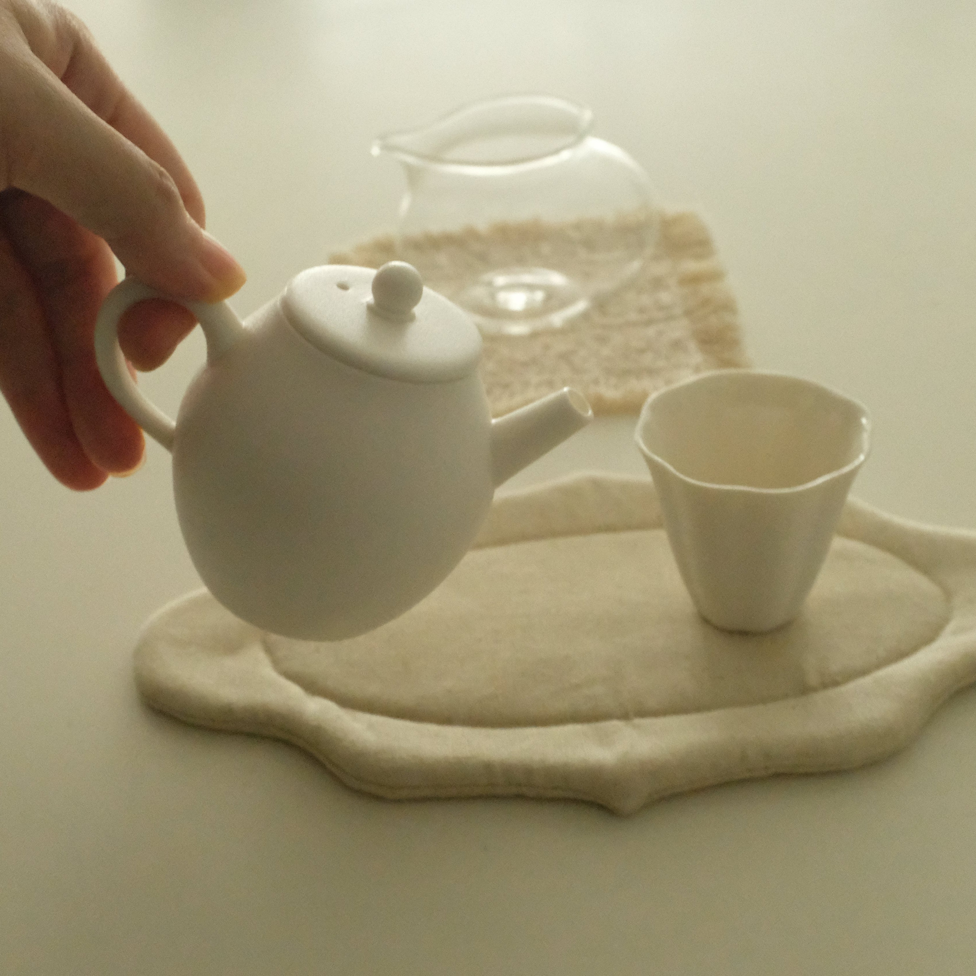 A hand holds a small white ceramic teapot above a matching white cup and a beige fabric tea mat on a light surface.