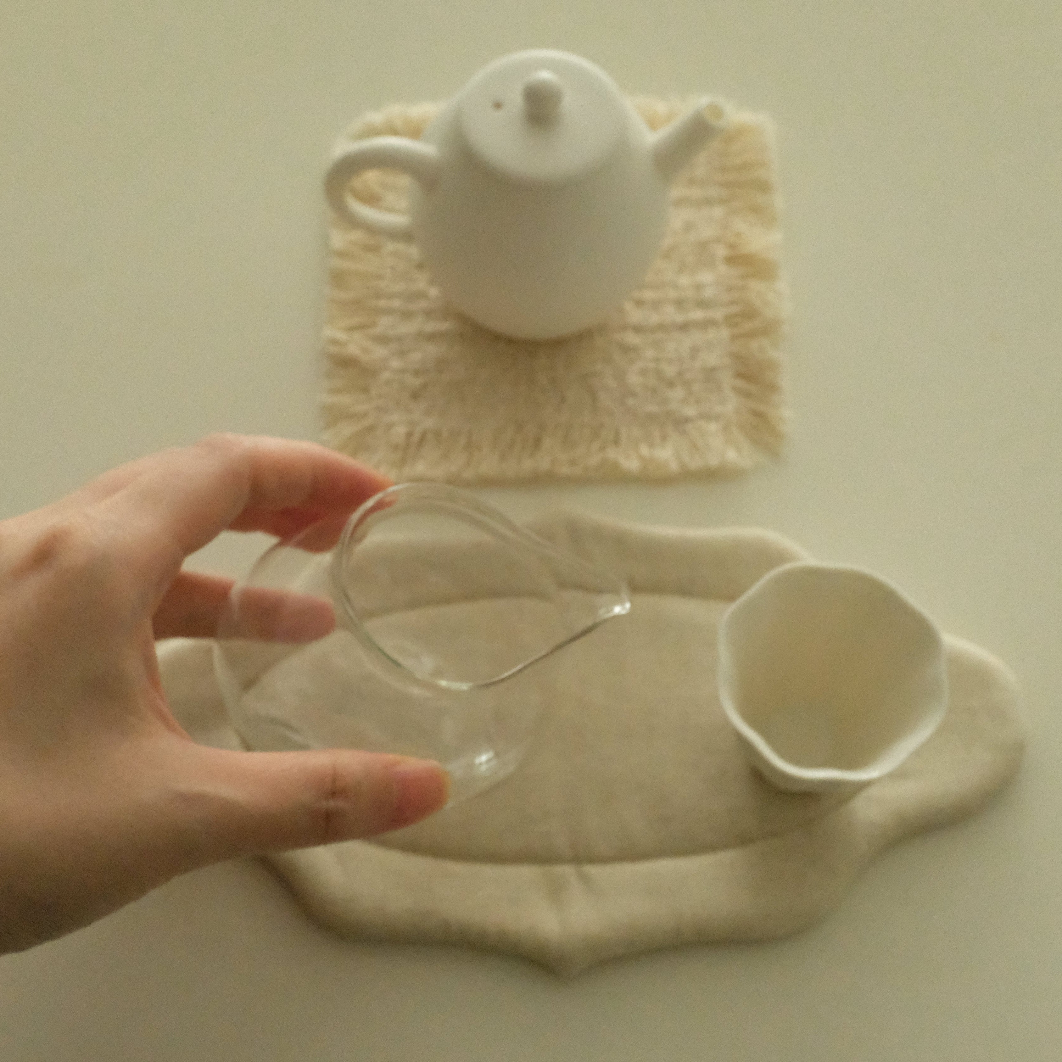 A hand holds a clear glass pitcher above a cream-colored fabric tea mat, with a white ceramic teapot and cup on matching mats in the background.