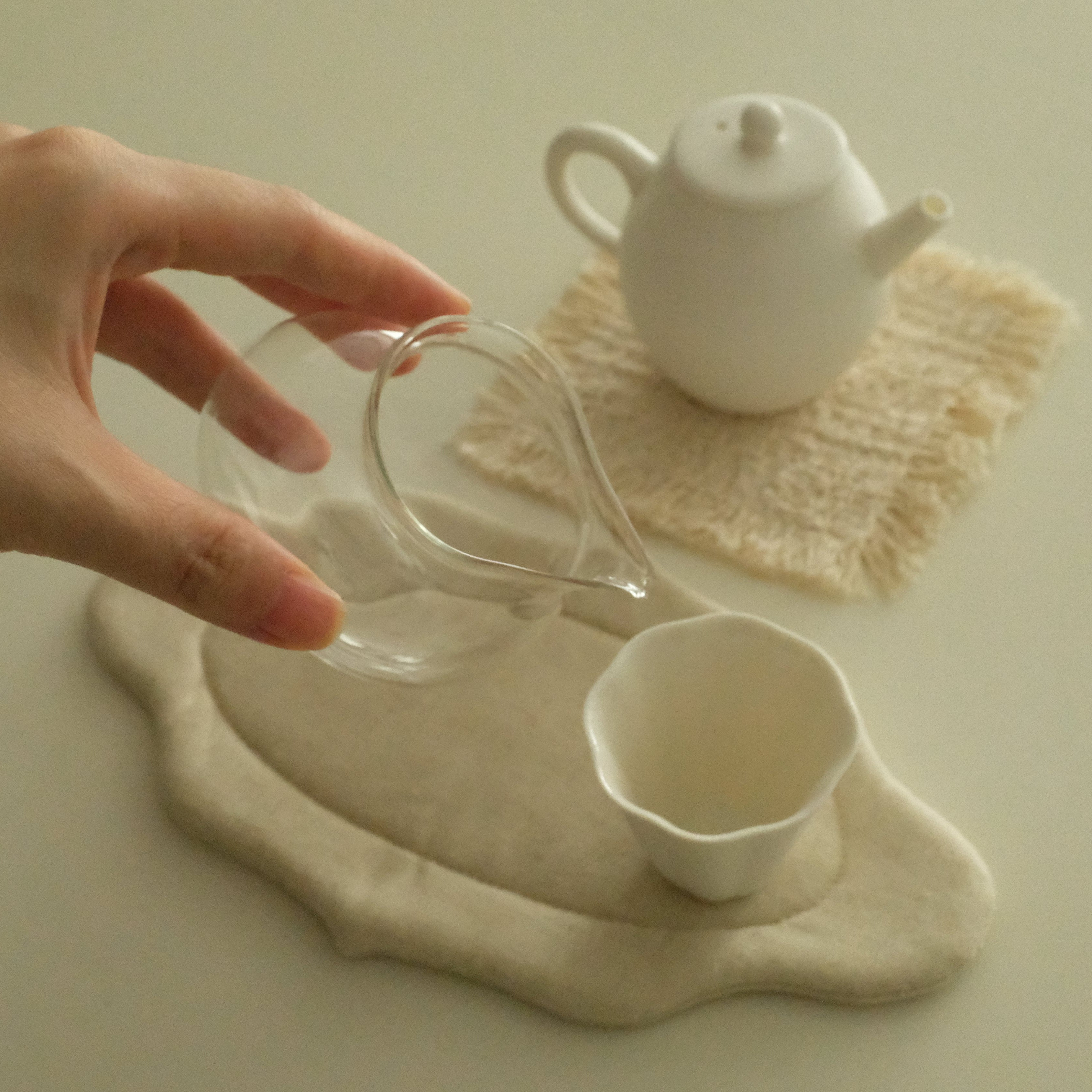 A hand holds a small clear glass pitcher above a white ceramic cup and a white teapot, arranged on a light beige surface with cream-colored mats.