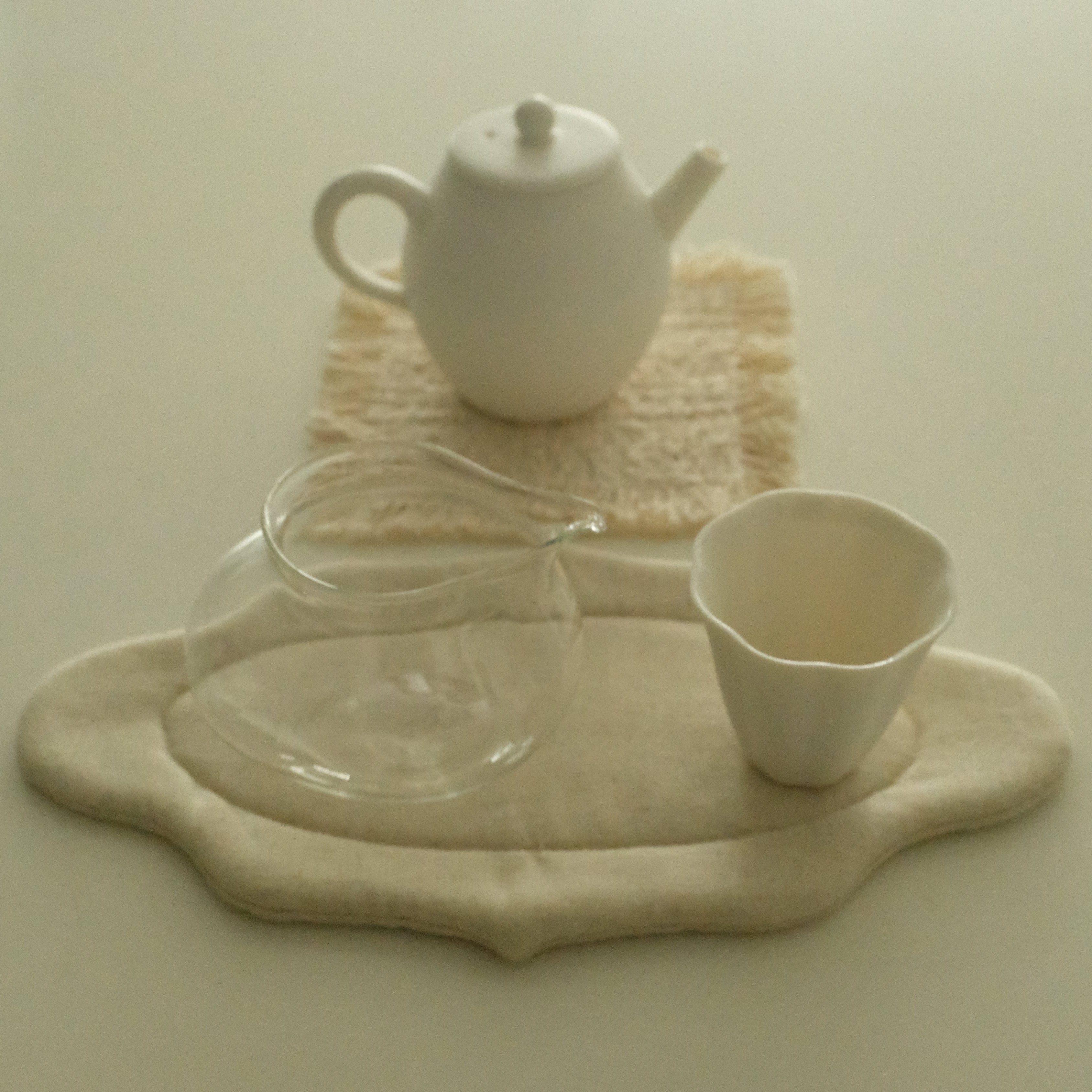 A minimalist tea set with a white ceramic teapot, a small white cup, and a clear glass fairness cup, arranged on cream-colored fabric placemats on a light beige surface.