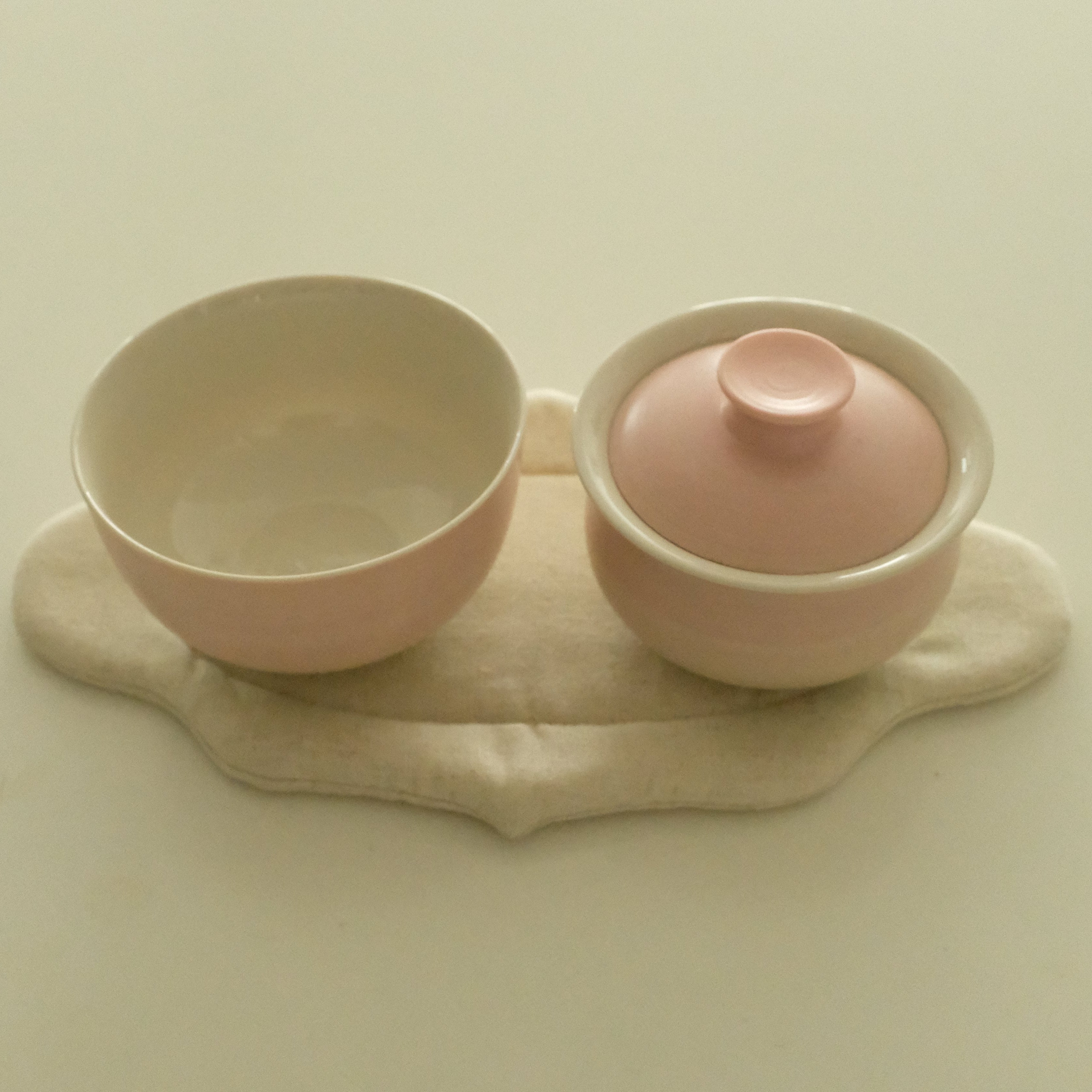 A pink ceramic tea set with a small lidded cup and matching saucer resting on a beige fabric pad, photographed against a plain cream background.