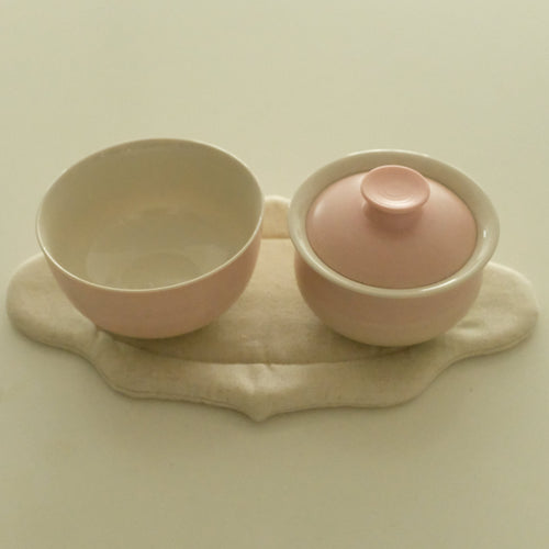 A pink ceramic tea set with a small lidded cup and matching saucer resting on a beige fabric pad, photographed against a plain cream background.