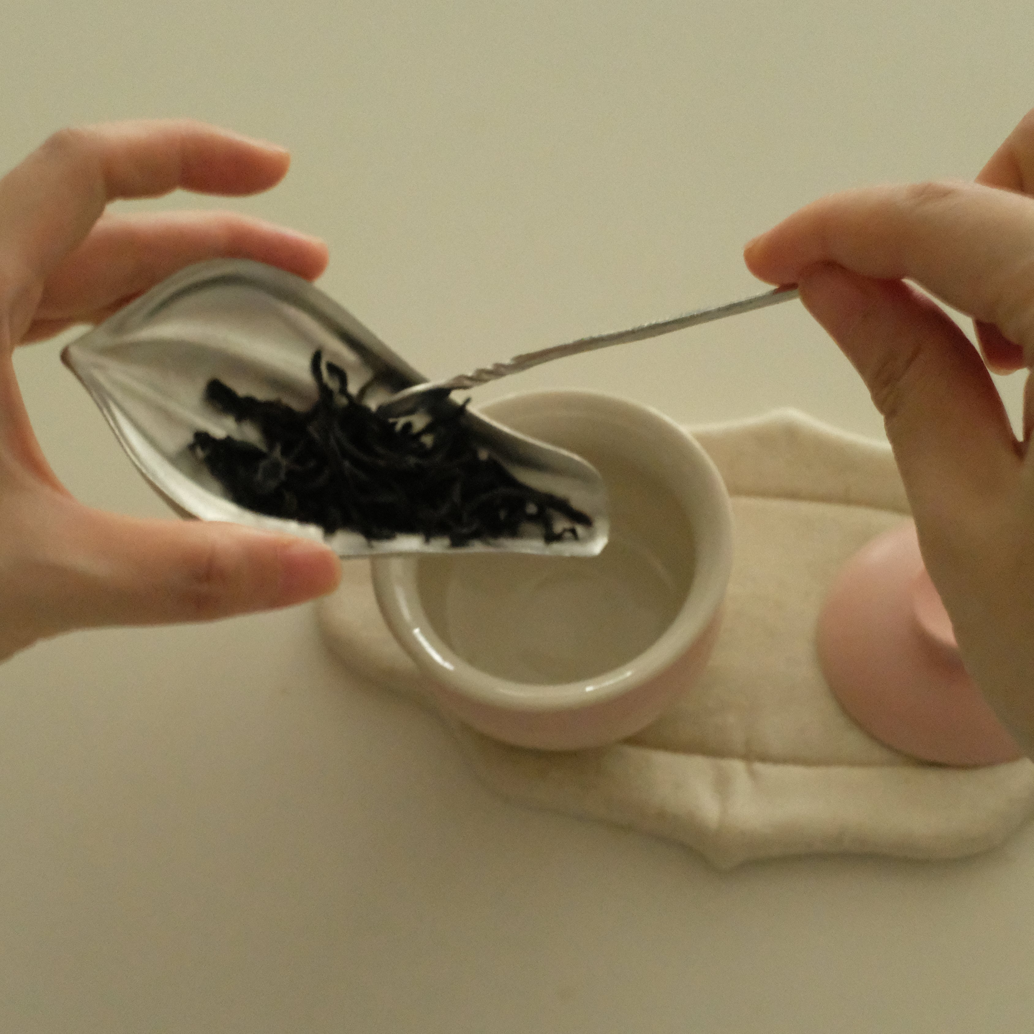 A close-up of hands preparing tea, pouring dark loose-leaf tea from a silver leaf-shaped scoop into a small white bowl, with a pink lid resting on a beige cloth in the background.