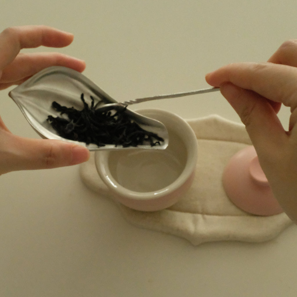 A close-up of hands holding a small silver leaf-shaped scoop filled with dark dried tea leaves above a small white cup and a pink lid on a cream cloth, set against a plain light background.