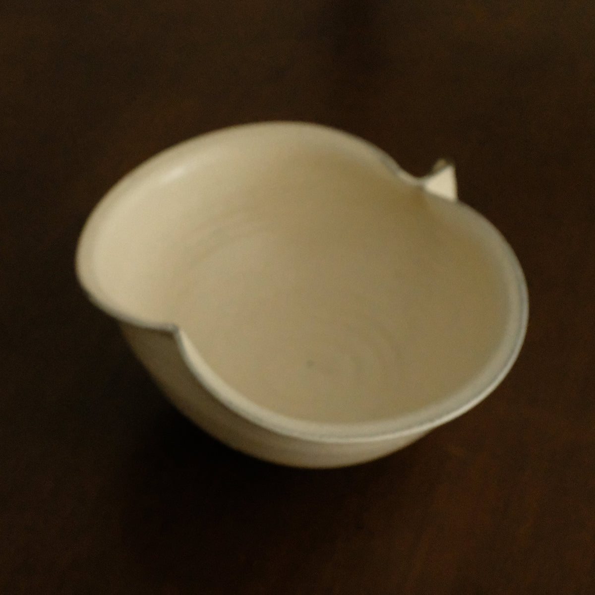 A small off-white ceramic bowl with a small tab-like protrusion on one side, photographed on a dark brown wooden surface.