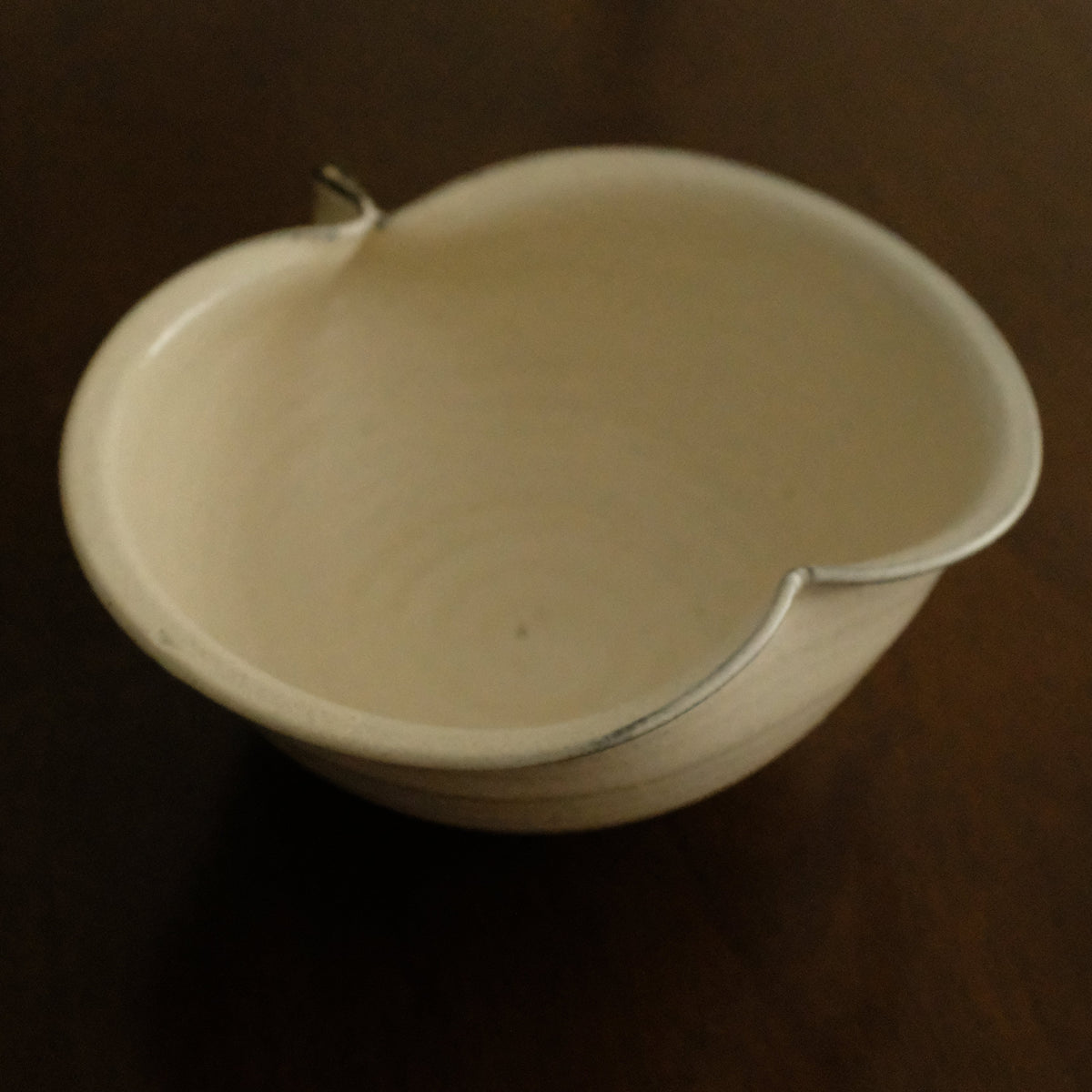 A small off-white ceramic bowl with a small tab-like protrusion at the top, photographed on a dark brown wooden surface.