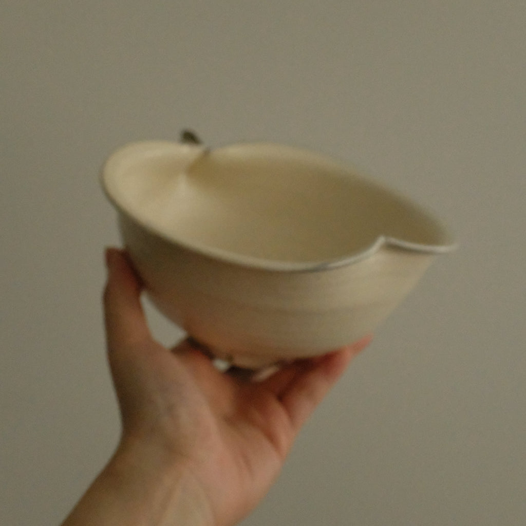A hand holding a small off-white ceramic bowl with a small tab-like detail on the rim against a plain gray background.