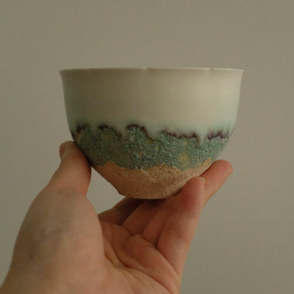 A handmade ceramic cup with a white glazed top half and a textured, speckled turquoise and brown bottom half, held in a person's hand against a plain beige background.