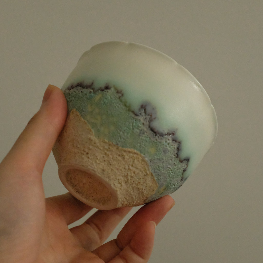 A hand holding a handmade ceramic bowl with a glossy white top section, a textured light green middle band, and a natural beige clay base.