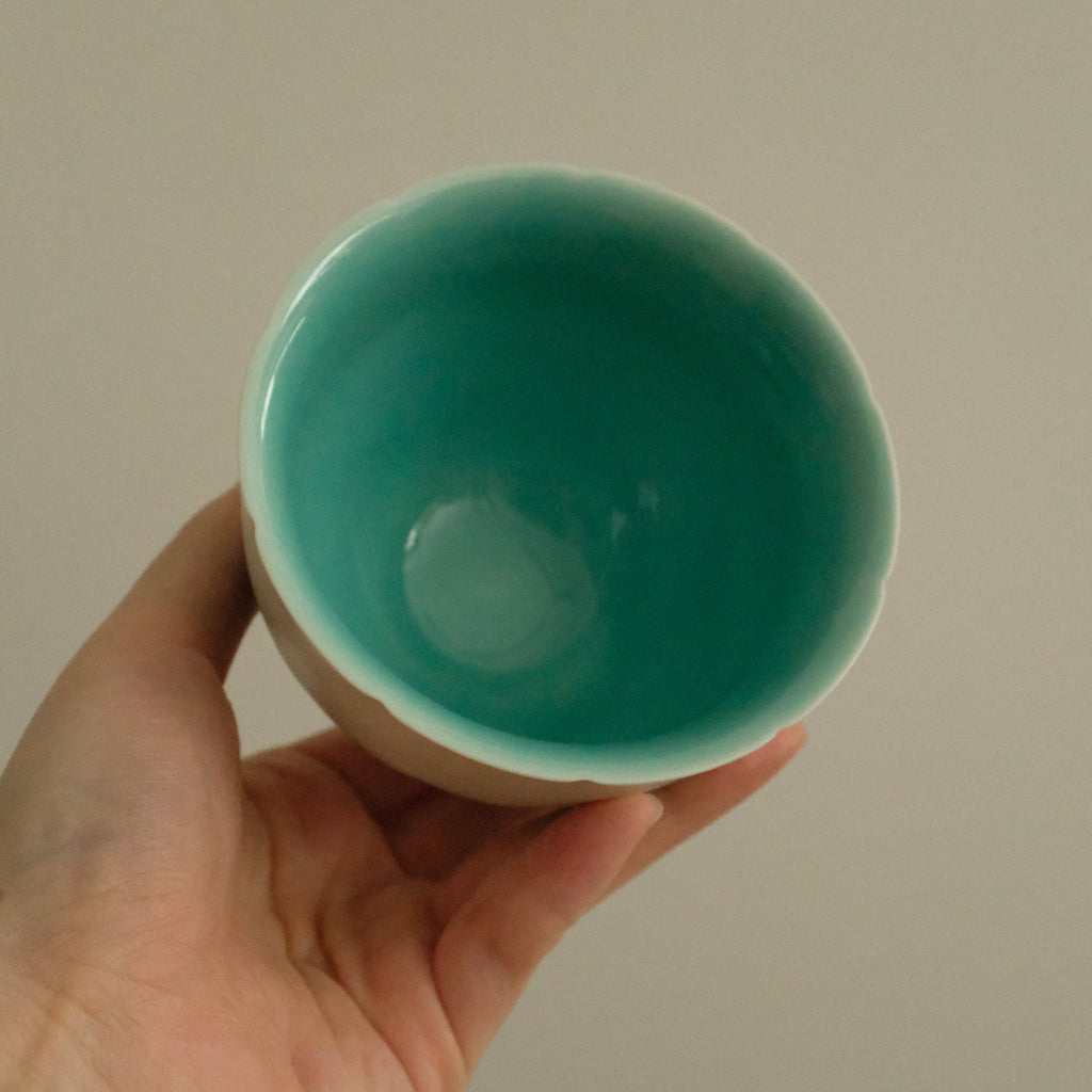A hand holding a small ceramic cup with a bright turquoise interior and a beige exterior, set against a plain light gray background.