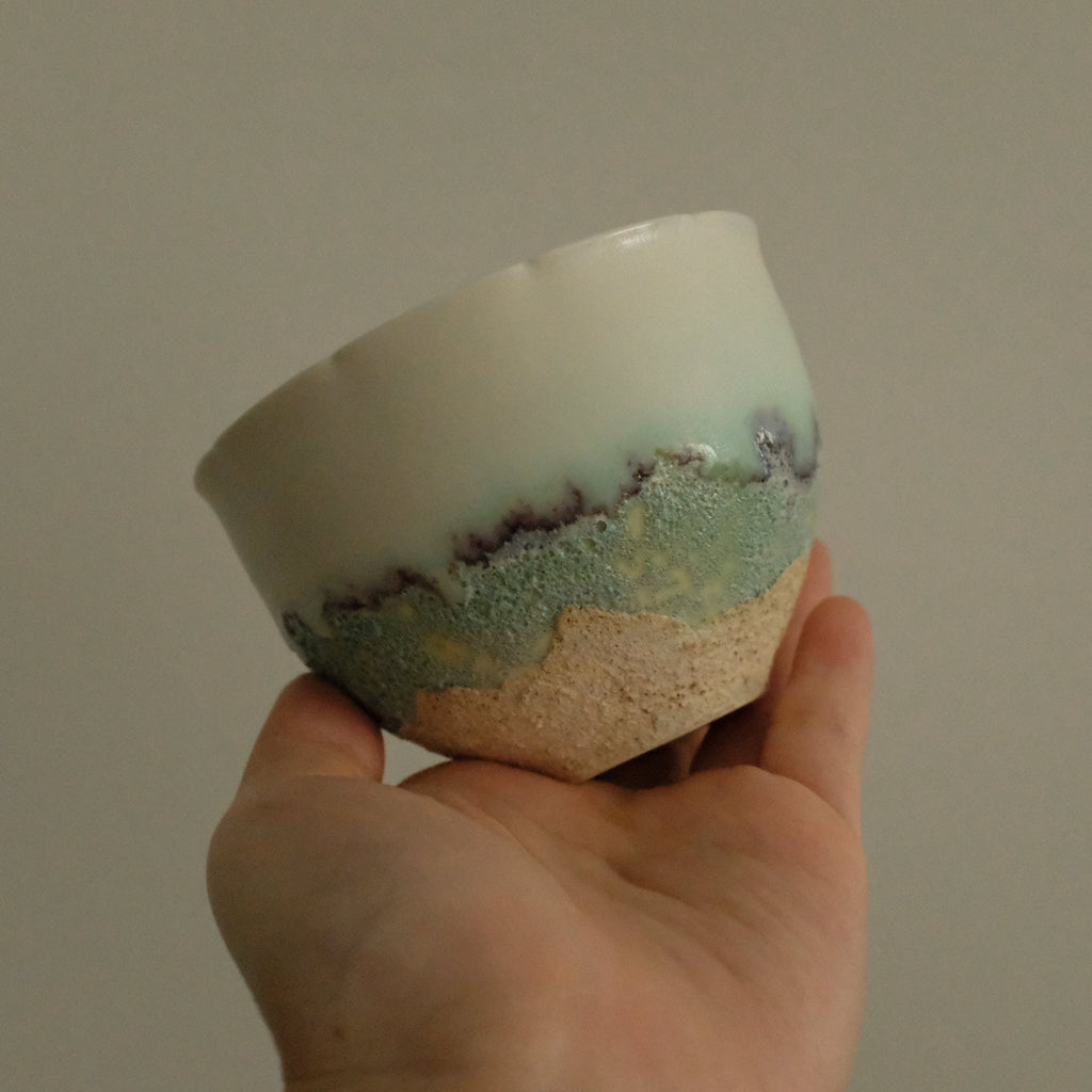 A handmade ceramic tea bowl with a white glazed top half, a textured green and blue middle band, and a rough beige clay base, held in a person's hand against a plain gray background.