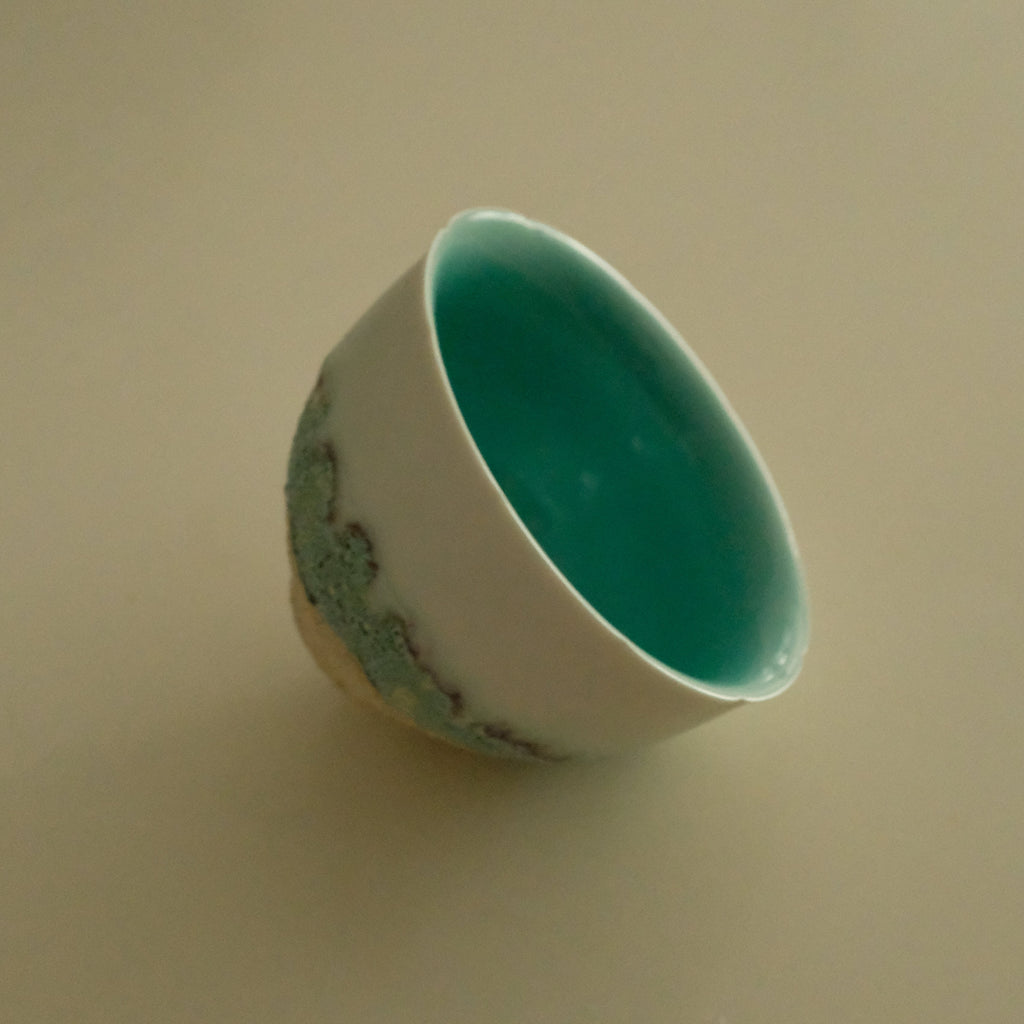 A small handmade ceramic cup with a glossy turquoise interior and a white exterior decorated with a green landscape pattern, shown on a plain beige background.