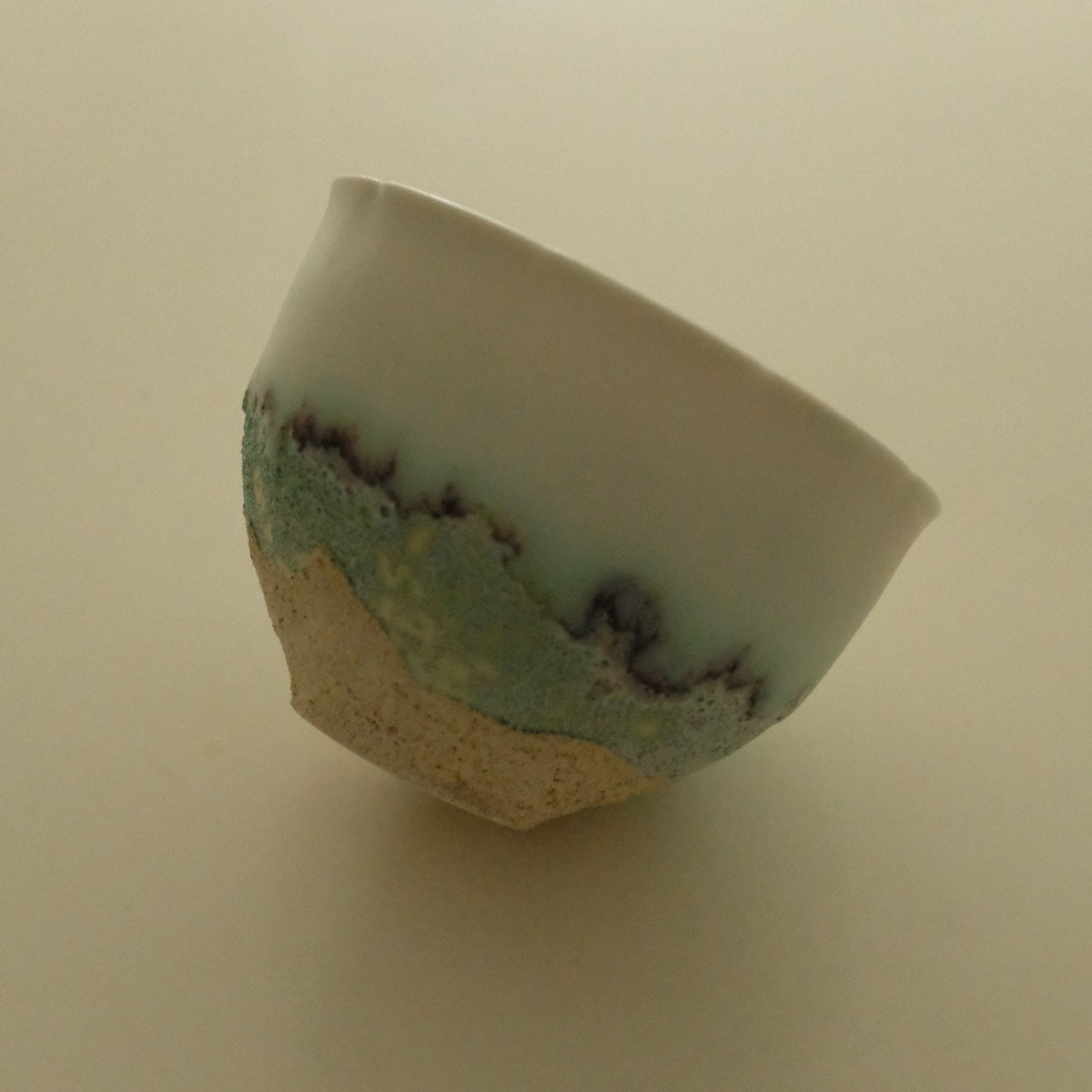 A handmade ceramic bowl with a glossy white upper half, a textured light blue lower half, and a natural beige unglazed base, photographed on a plain beige background.