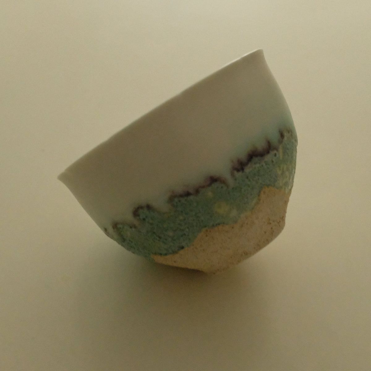 A handmade ceramic bowl with a white glazed upper half, a textured turquoise-green lower band, and a natural beige clay base, photographed on a plain beige background.