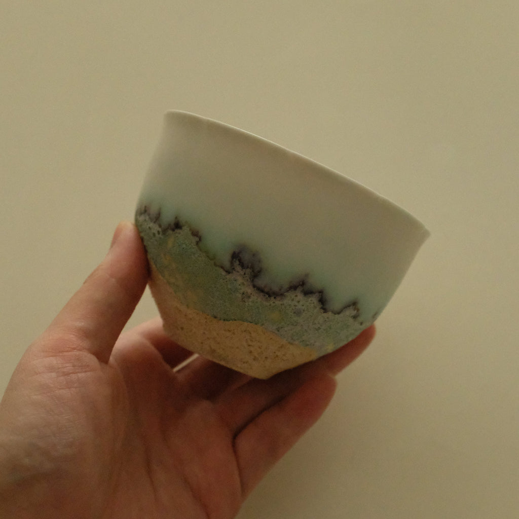 A hand holding a handmade ceramic bowl with a white glazed top half, a textured green band with dark speckles in the middle, and a natural beige clay base.