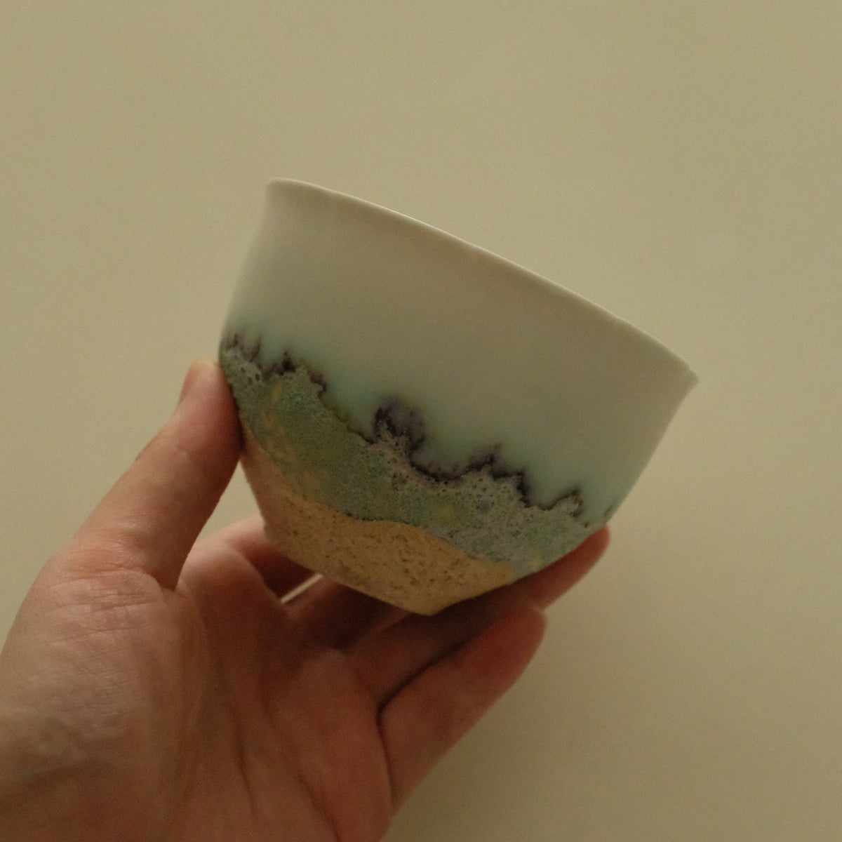 A hand holding a handmade ceramic bowl with a white glazed top half, a textured green band with dark speckles in the middle, and a natural beige clay base.
