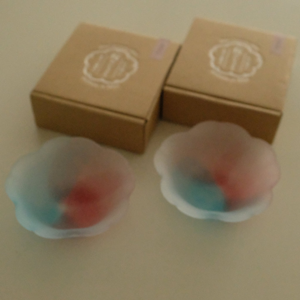 Two small translucent flower-shaped bowls containing red and blue contents, placed in front of two brown cardboard boxes with a circular logo on a beige surface.