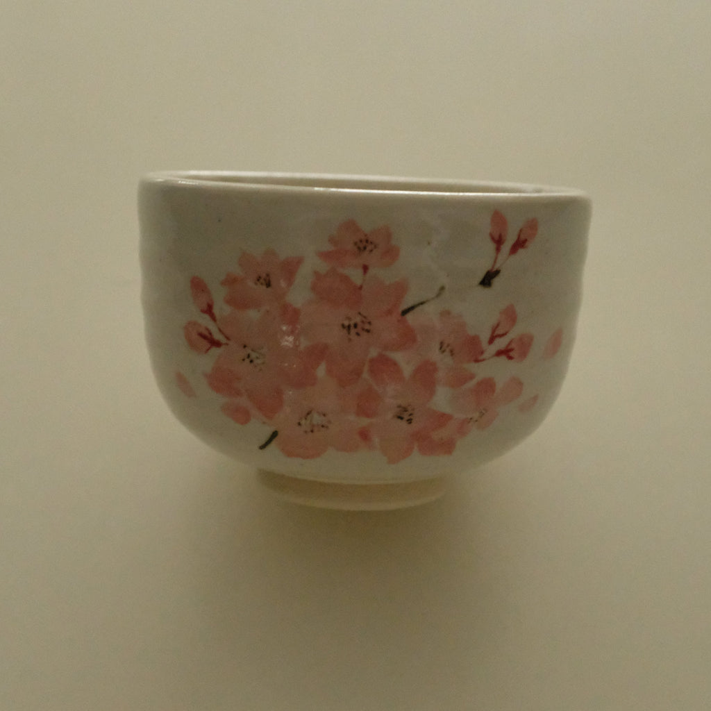A white ceramic bowl decorated with pink cherry blossom flowers and small black stems, set against a plain beige background.