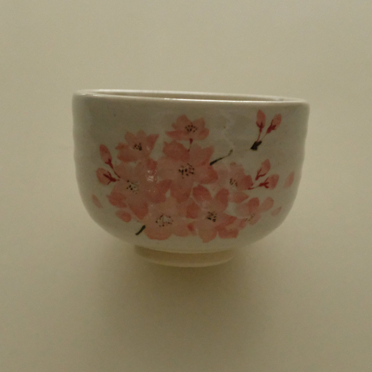 A white ceramic bowl decorated with pink cherry blossom flowers and small black stems, set against a plain beige background.