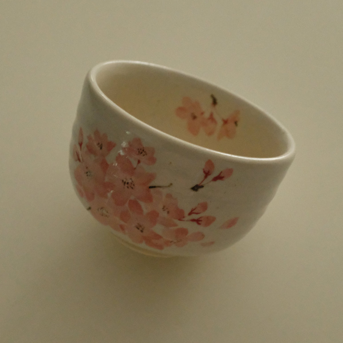 A small white ceramic cup decorated with pink cherry blossom flowers and dark stems, photographed against a plain beige background.