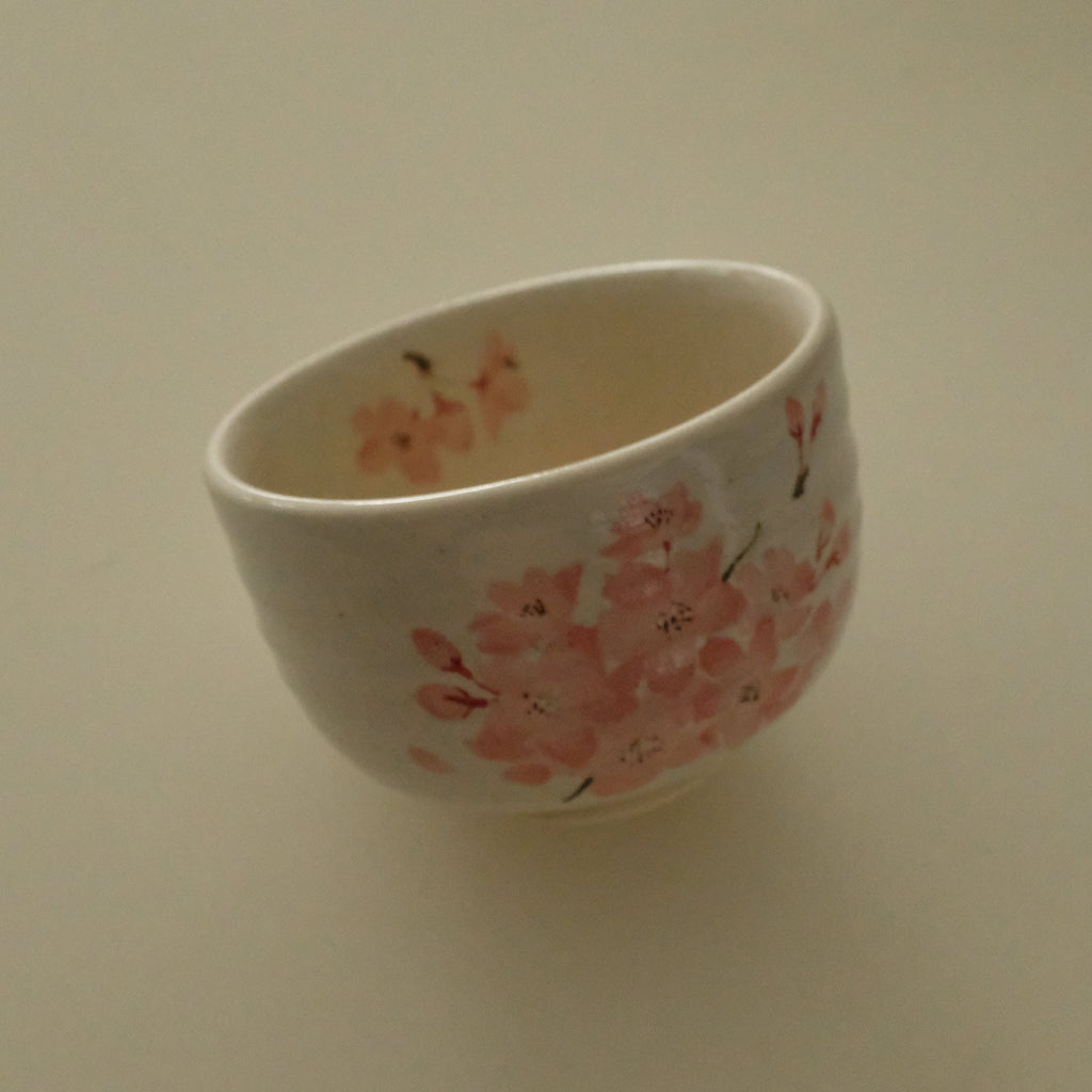 A small white ceramic cup decorated with pink cherry blossom patterns, shown on a plain beige background.
