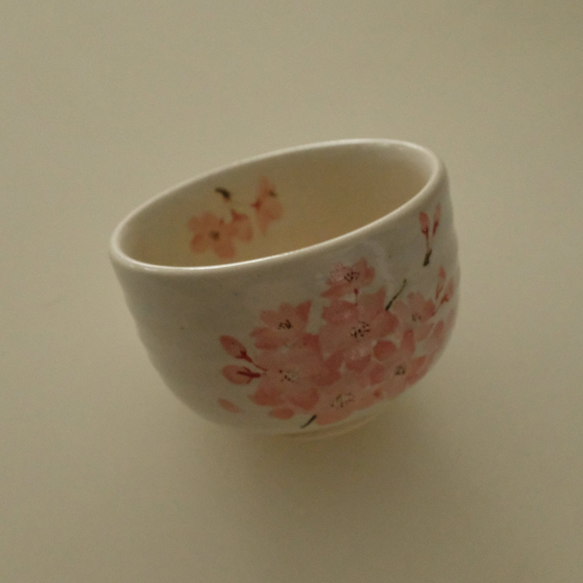 A small white ceramic cup decorated with pink cherry blossom patterns, shown on a plain beige background.