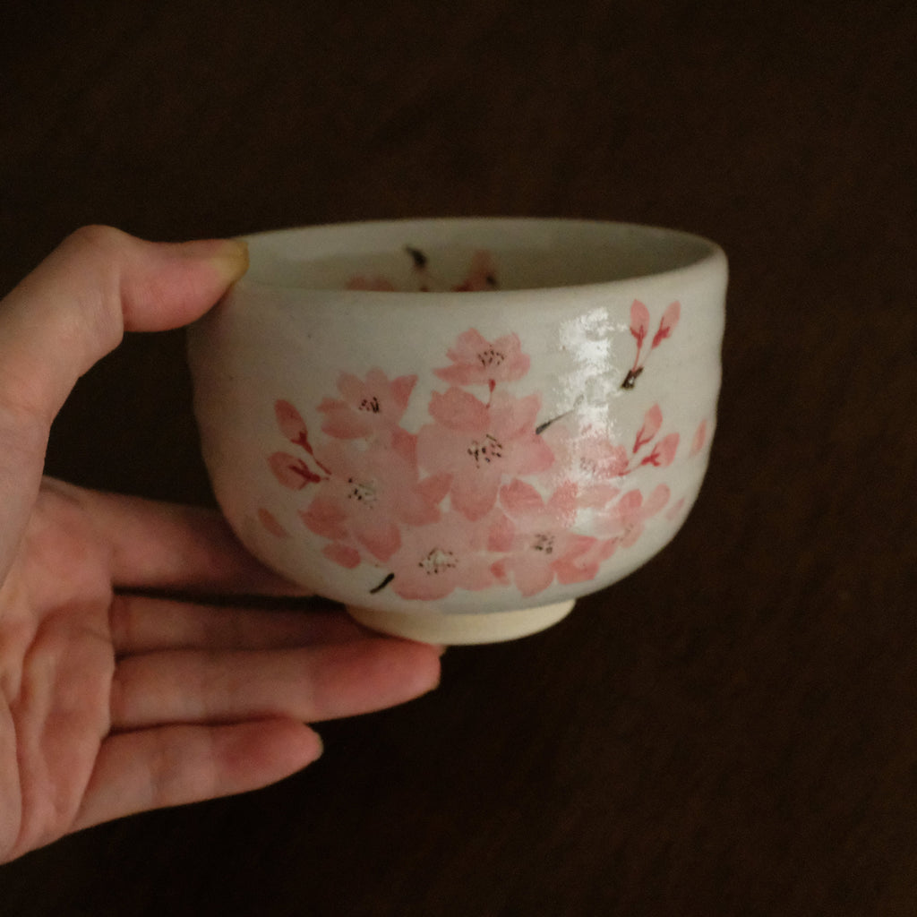 A hand holding a small white ceramic bowl decorated with pink cherry blossom flowers, set against a dark brown background.