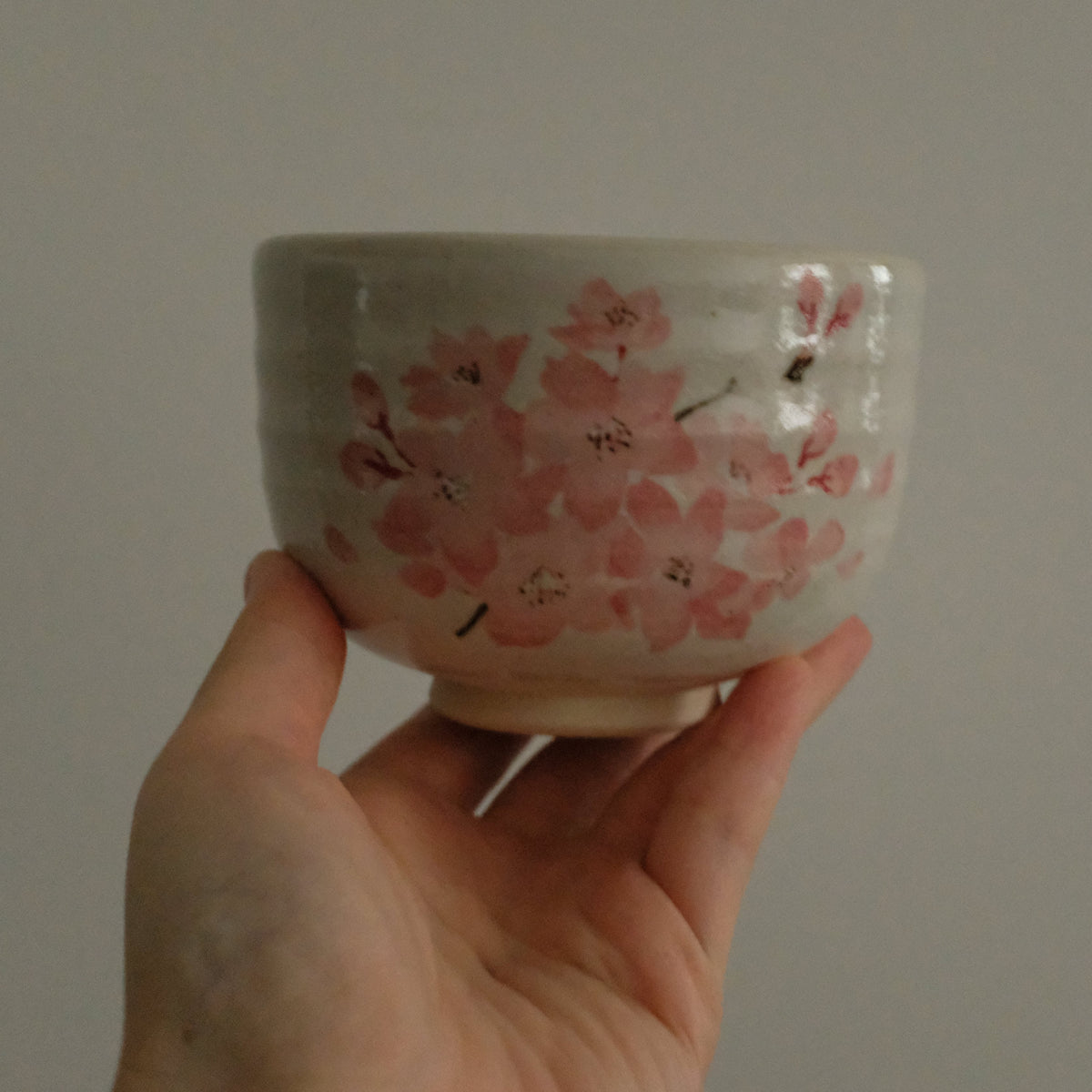 A hand holding a small ceramic bowl with a pink cherry blossom pattern on a light gray background.