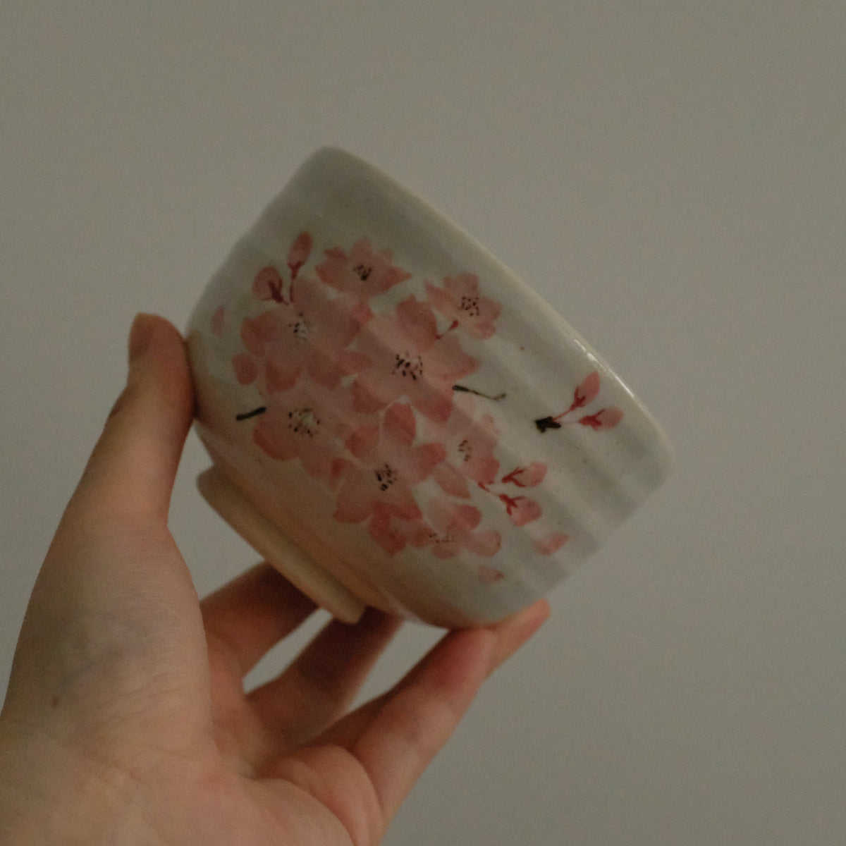A hand holding a small white ceramic bowl decorated with pink cherry blossom flowers and small red insects, set against a plain light gray background.