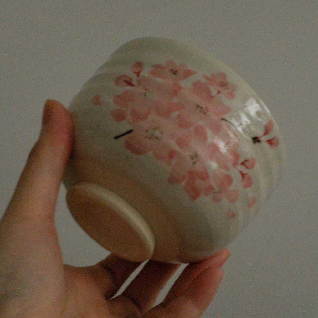 A hand holding a small white ceramic bowl decorated with pink cherry blossoms, set against a plain gray background.