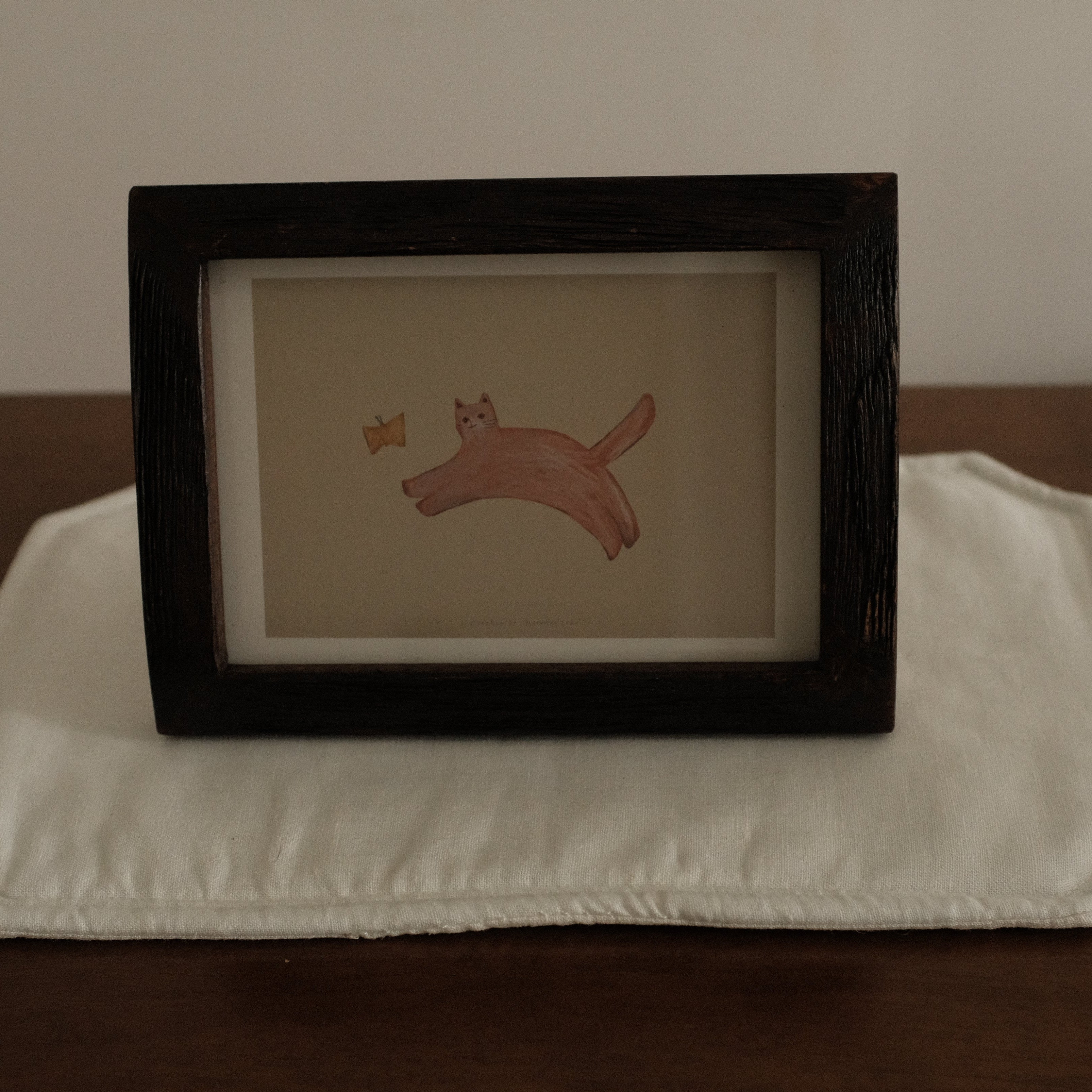 Framed artwork of a cat with butterfly on a white placemat with a brown background