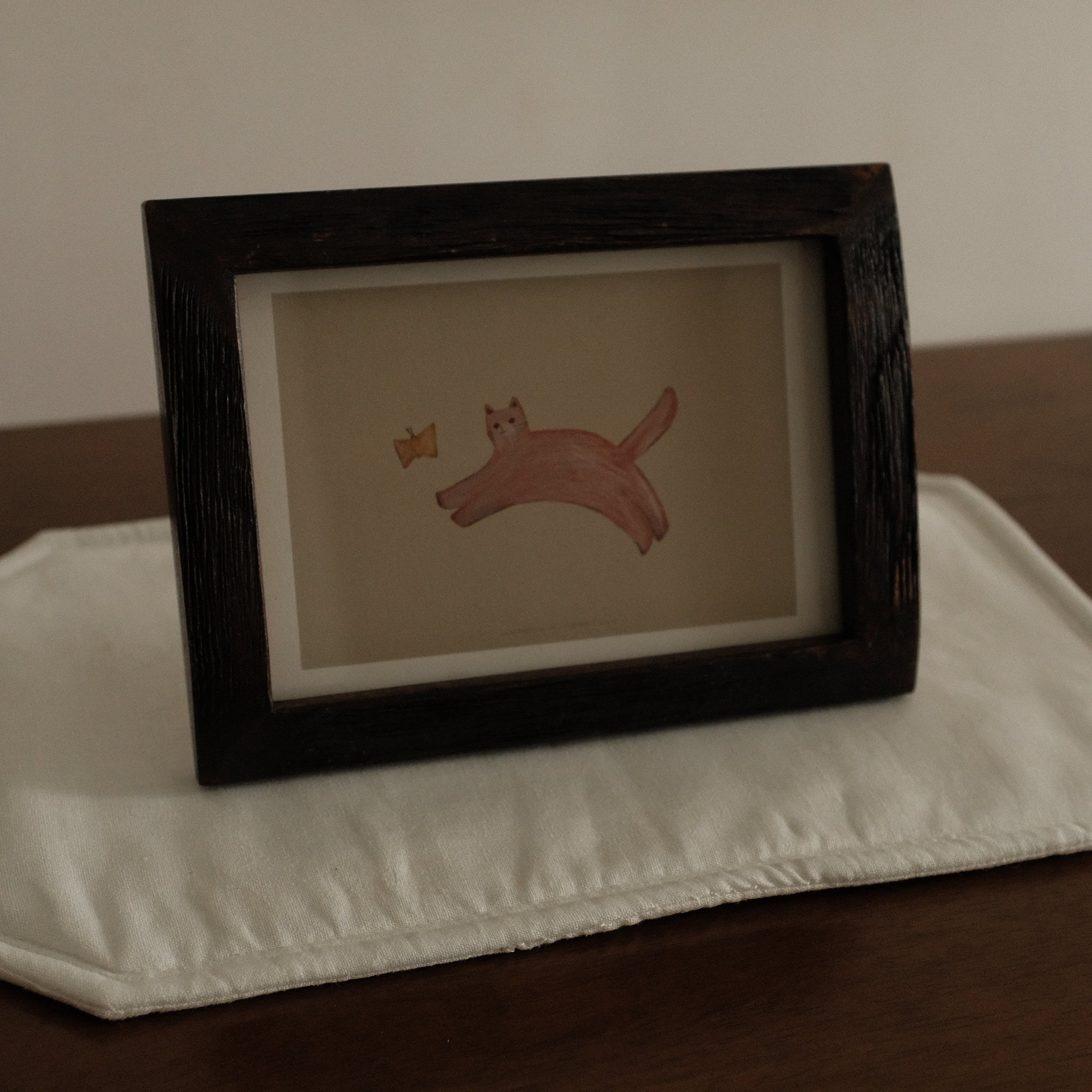 Framed artwork of a cat with butterfly on a white placemat with a brown background