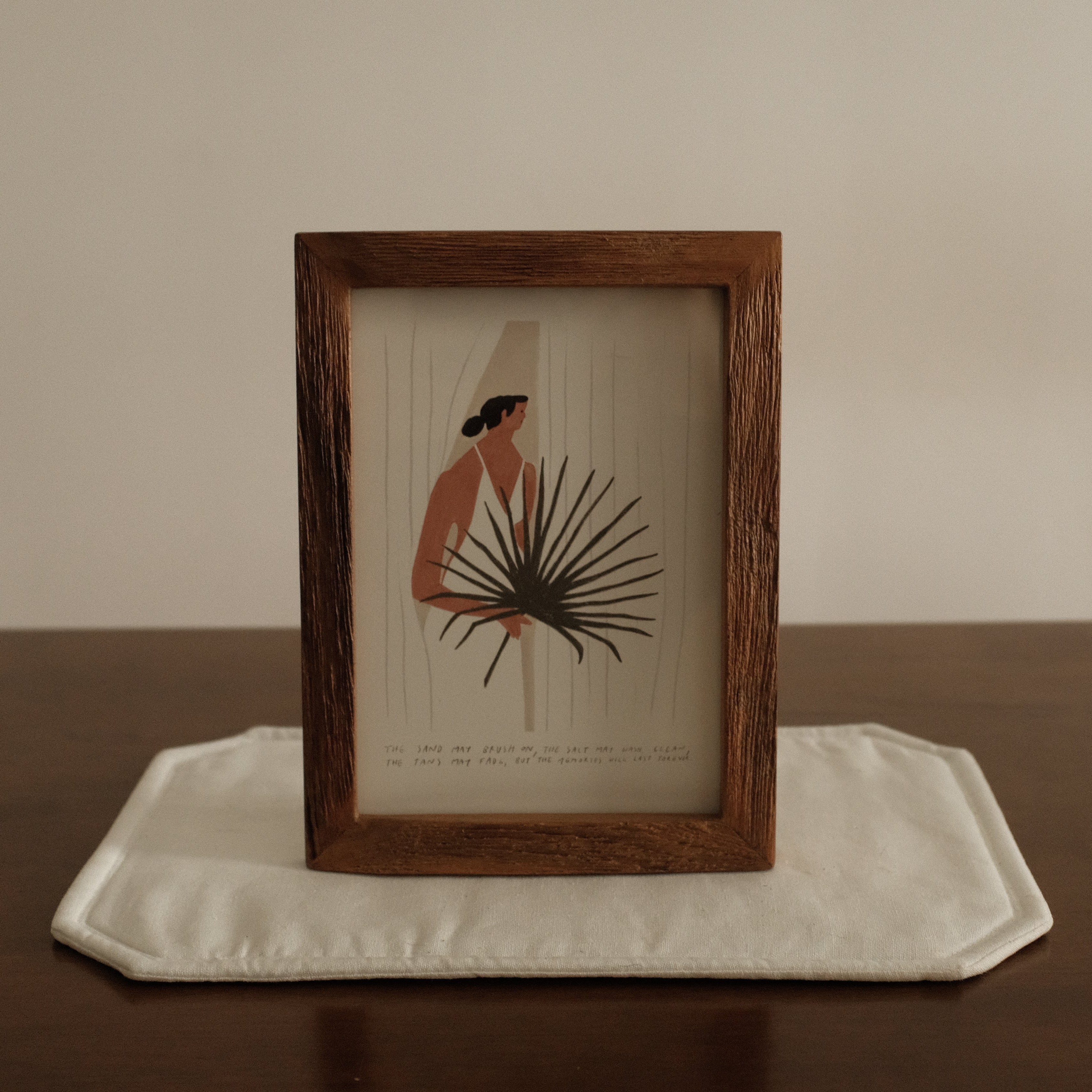 Framed artwork of a person with a plant on a white tray against a beige wall.