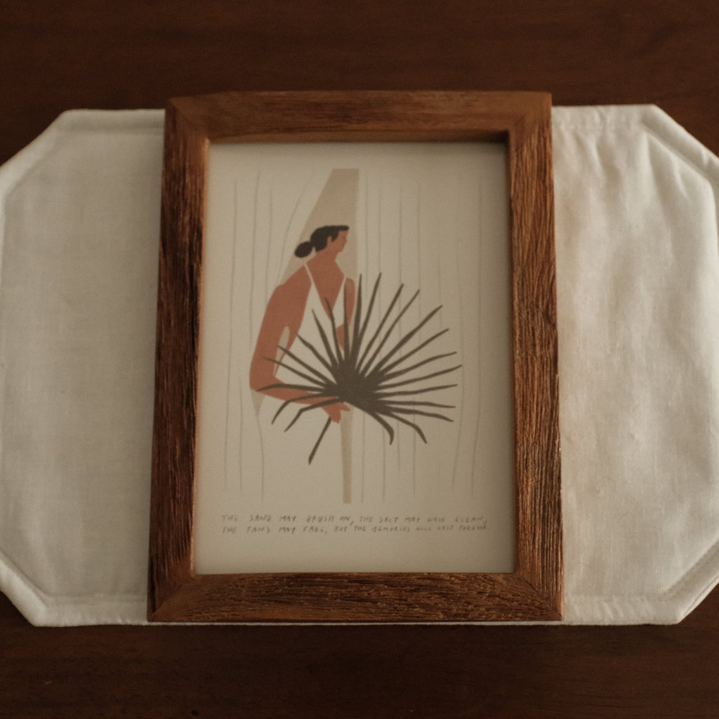 Framed artwork of a woman holding a palm leaf on a white surface with a brown background