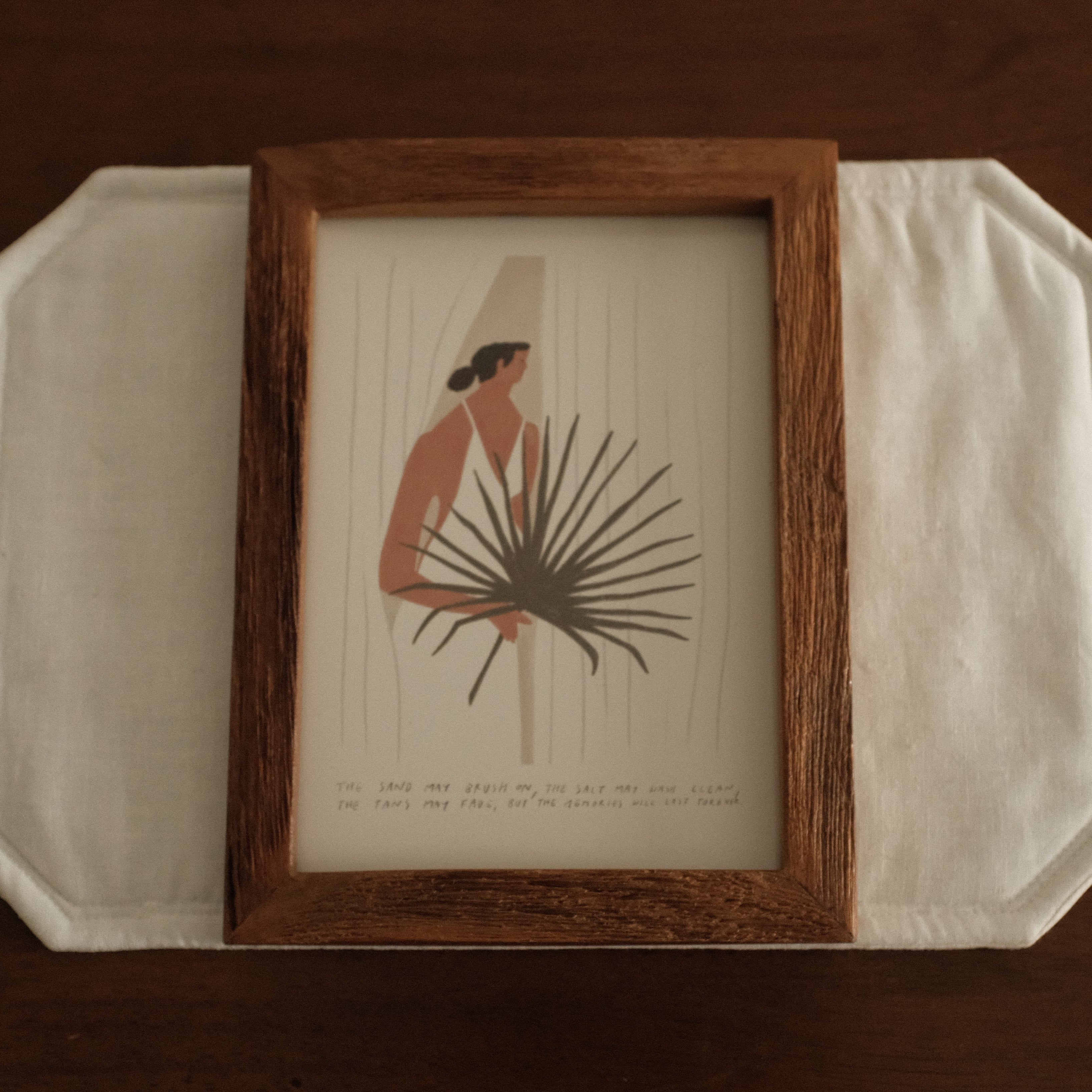 Framed artwork of a woman holding a palm leaf on a white surface with a brown background