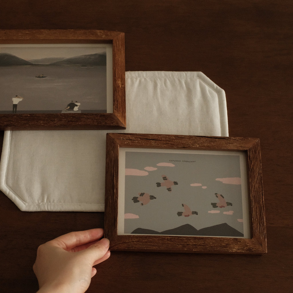Two teak wooden-framed artworks on a dark surface with a hand reaching towards one of them.