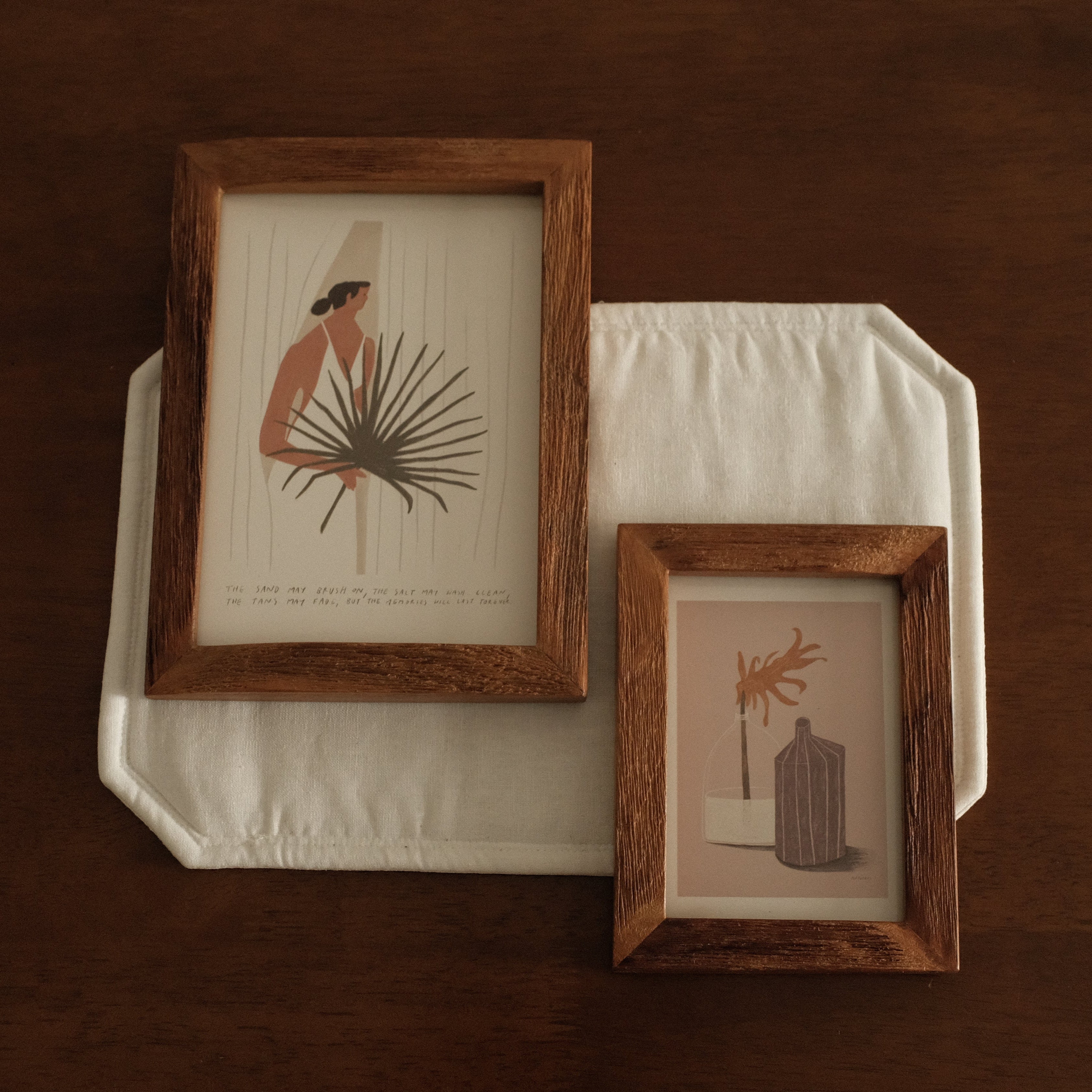 Two wooden picture frames with prints on a white cloth against a brown background
