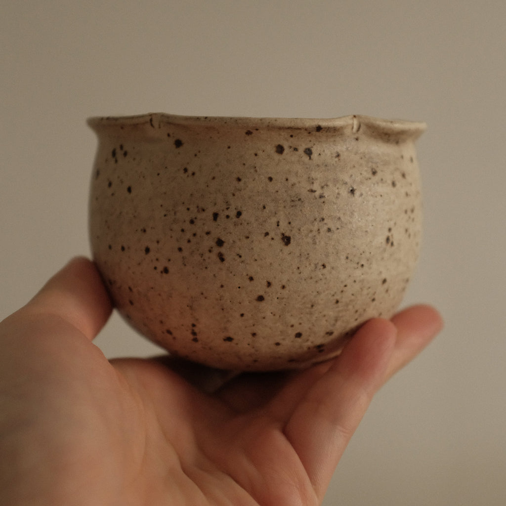 Hand holding a small speckled ceramic grey beige floral rim cup against a plain background