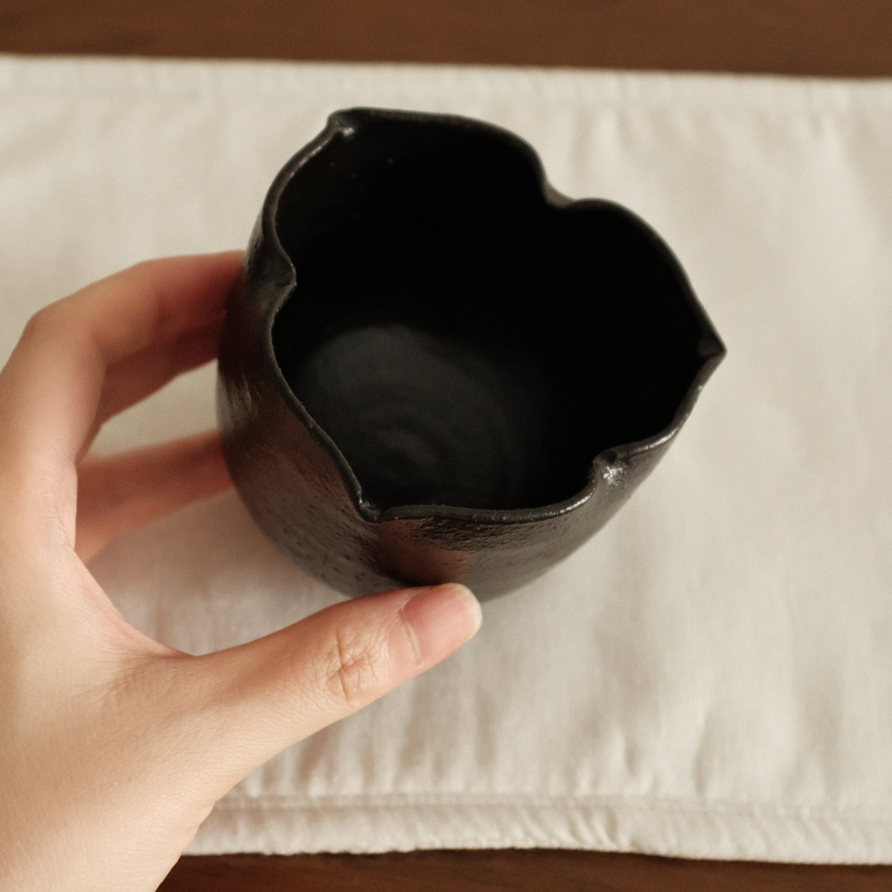 Hand holding a black three petals ceramic cup on a white square mat with a brown background