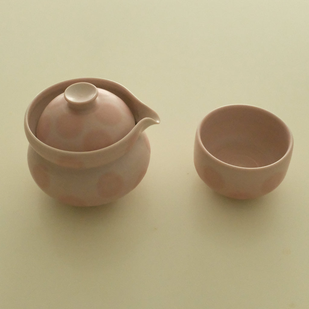 A small pink ceramic teapot with a lid and spout, shown next to a matching pink cup on a plain cream-colored background.