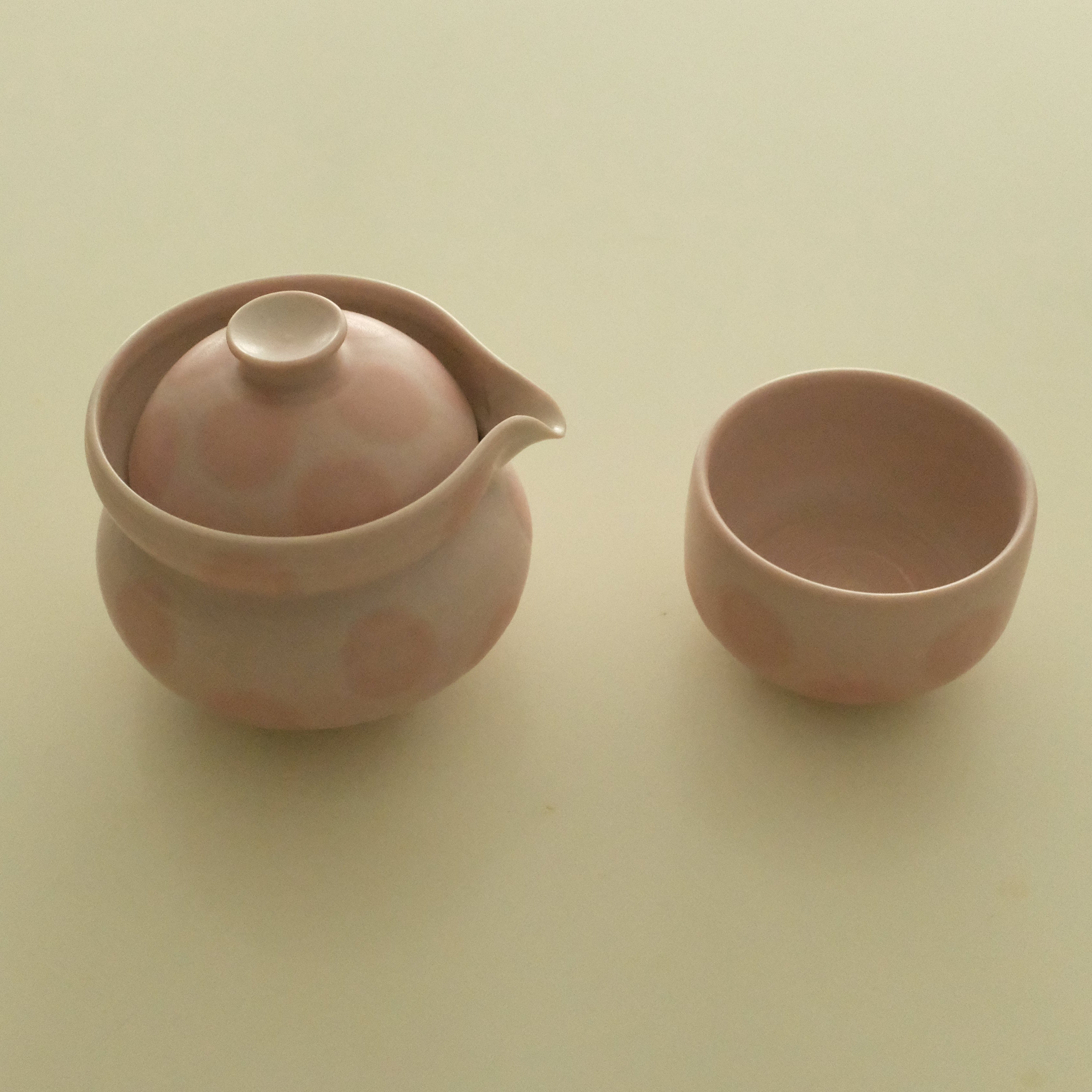 A small pink ceramic teapot with a lid and spout, shown next to a matching pink cup on a plain cream-colored background.