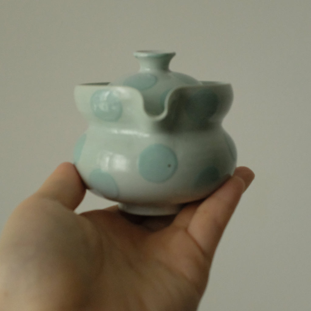 A hand holding a small ceramic teapot with a pale green glaze and light blue polka dots against a plain light gray background.