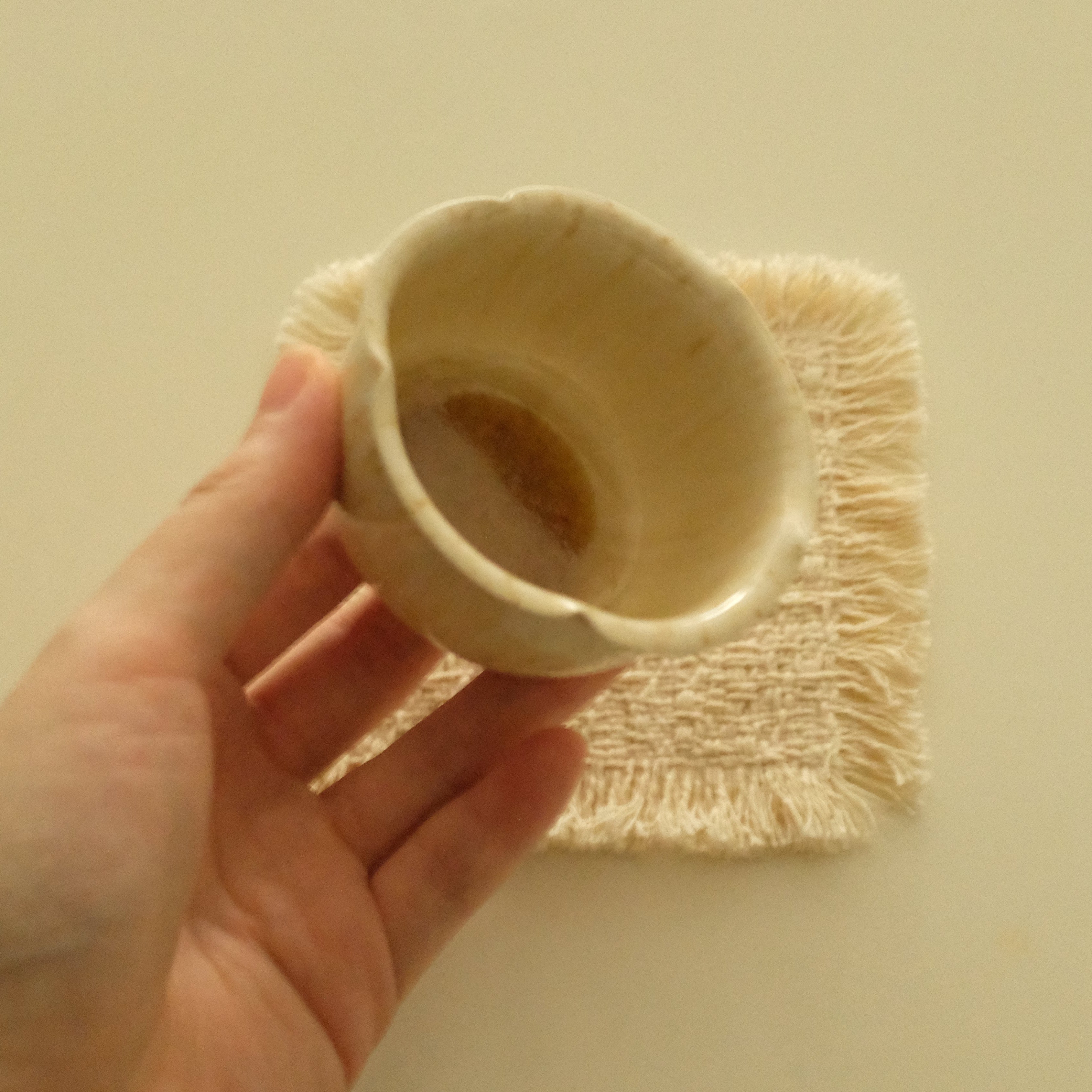 A hand holding a small handmade ceramic cup with a scalloped rim, set against a plain beige background with a small woven napkin underneath.