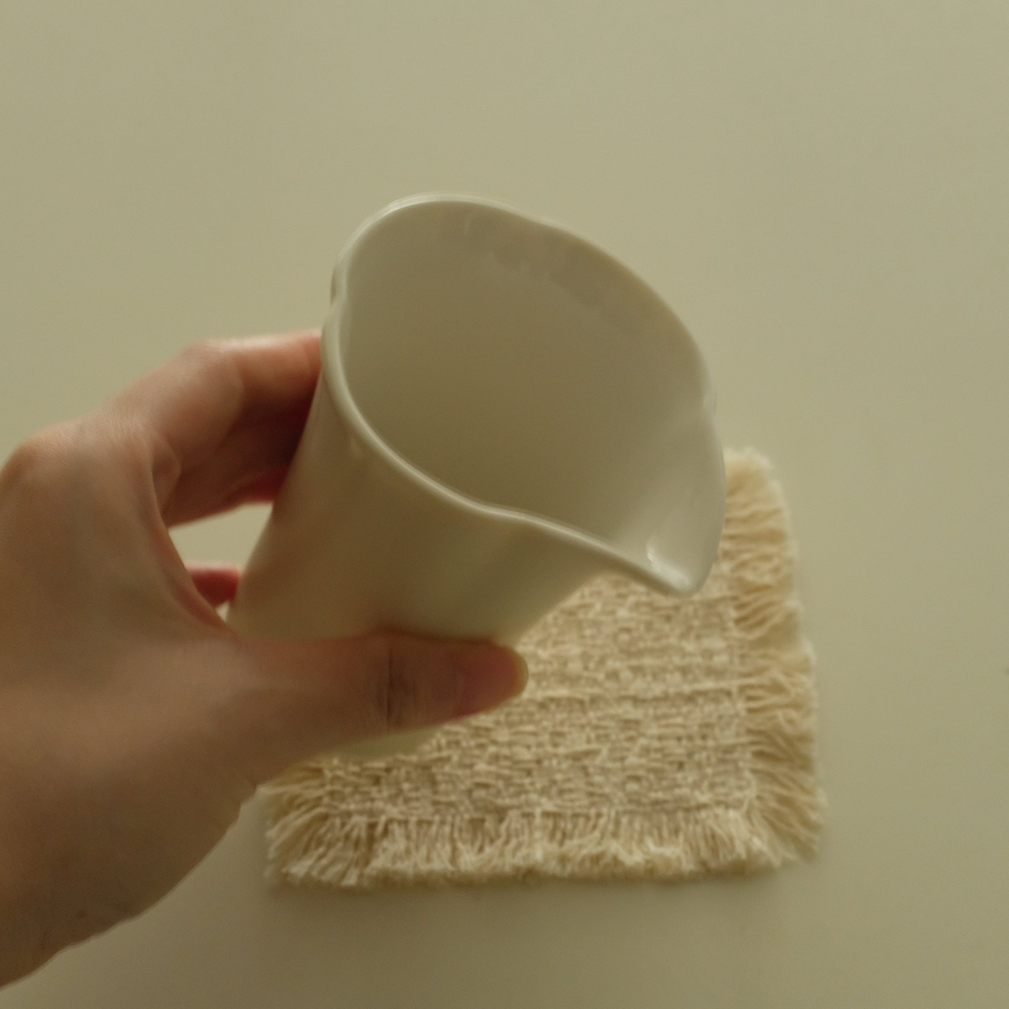 A hand holding a small off-white ceramic pitcher or cup with a pouring spout, positioned over a textured cream-colored cloth on a plain light beige background.