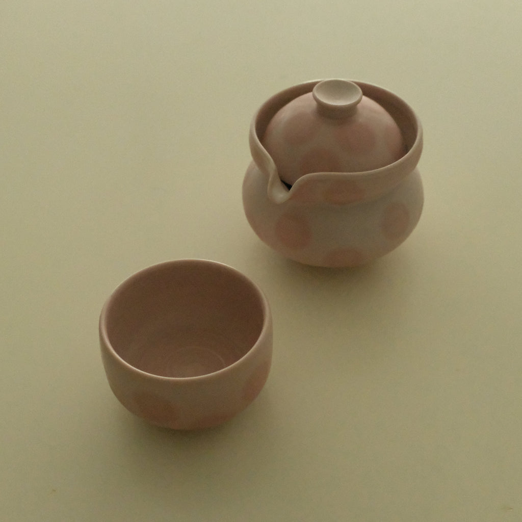 A small pink ceramic teapot with a matching lid and a separate small cup, photographed on a plain light beige background.